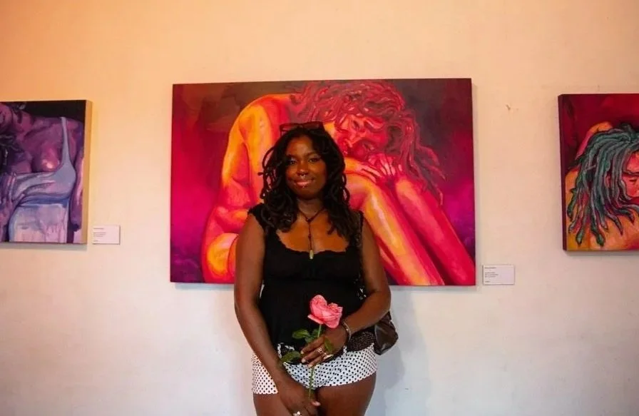 A woman holding a pink flower standing in front of colorful paintings featuring women in an art gallery.