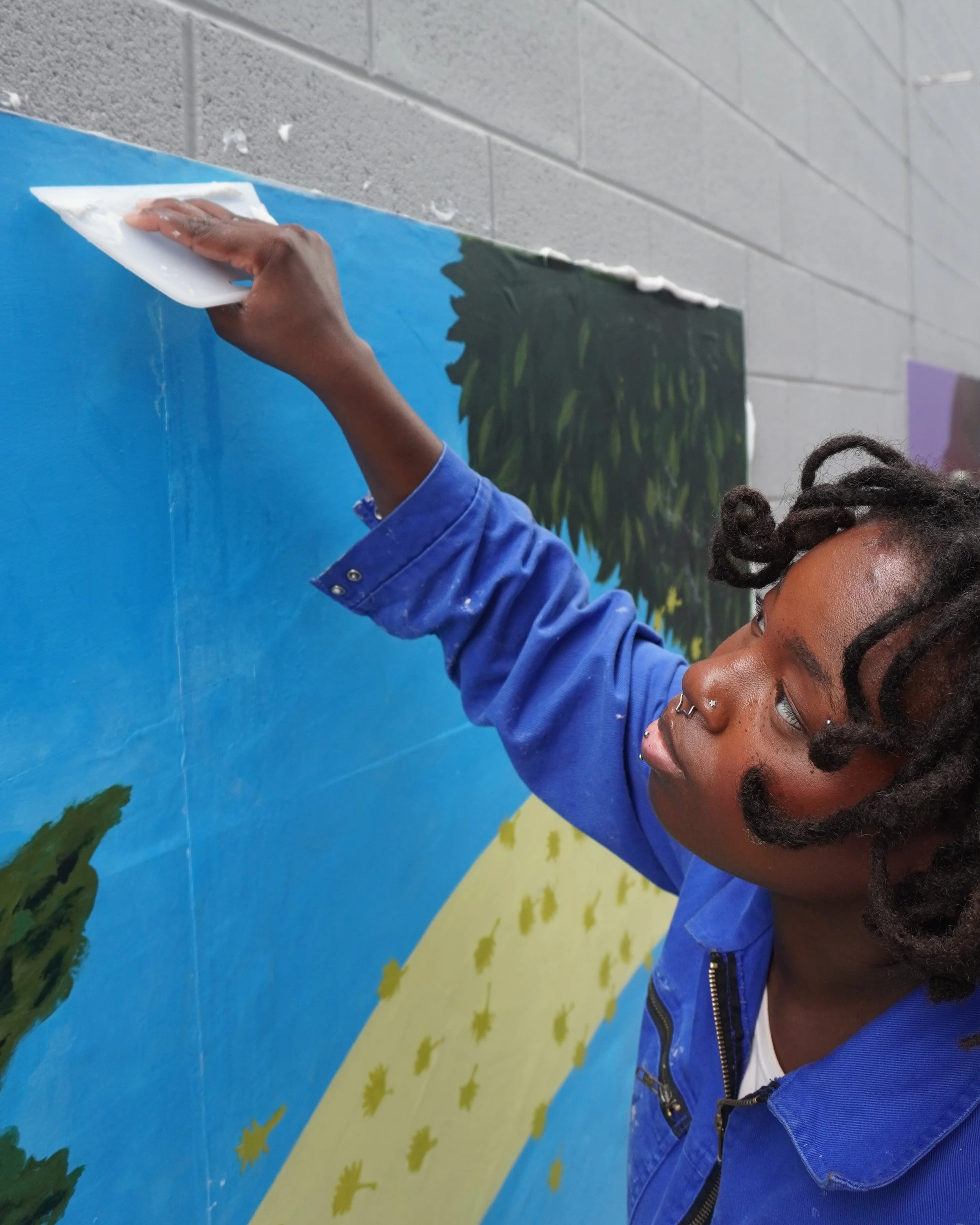 A person with dark skin and dreadlocks painting on a colorful mural with a sponge, wearing a blue jacket and a white shirt, in an indoor setting with a gray cinder block wall.