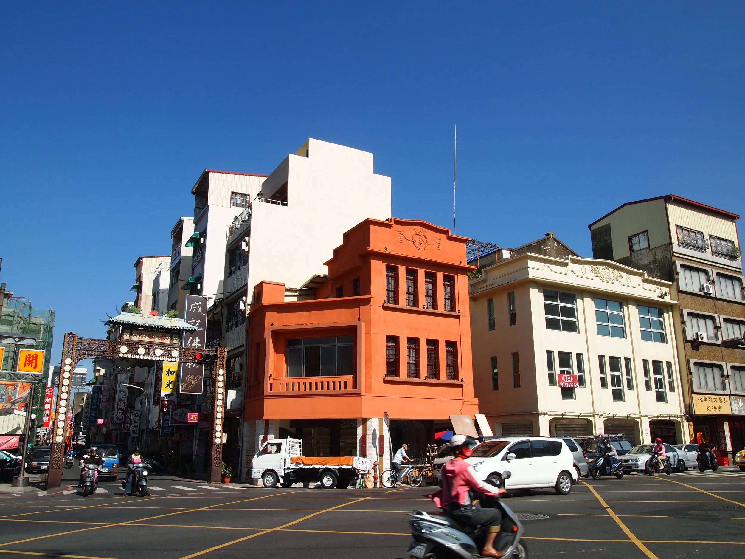 City street scene with colorful buildings, scooters, cars, and pedestrians under a bright blue sky.