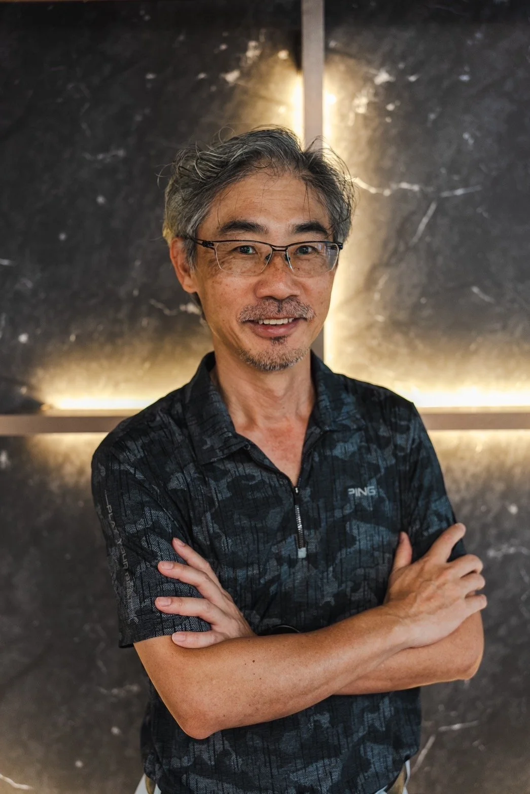 A middle-aged man with gray hair, glasses, and a goatee smiling with crossed arms, standing in front of a dark marble wall with backlighting.