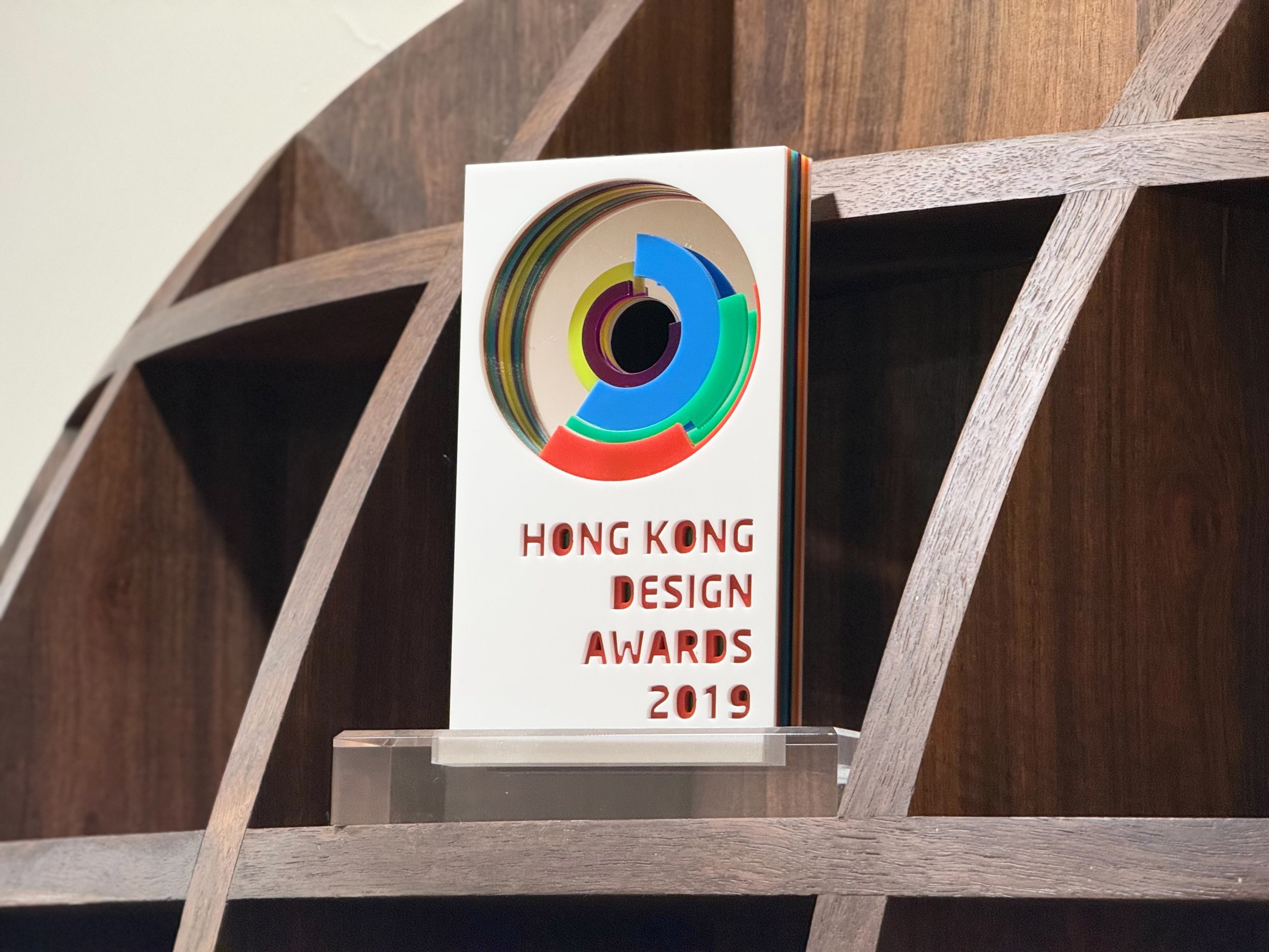 A clear acrylic award plaque featuring colorful concentric circular design element, with the text 'HONG KONG DESIGN AWARDS 2019' written on it, placed on a wooden surface.榮獲 2019 香港設計大獎 Hong Kong Design Awards 之台中建築師事務所獎牌