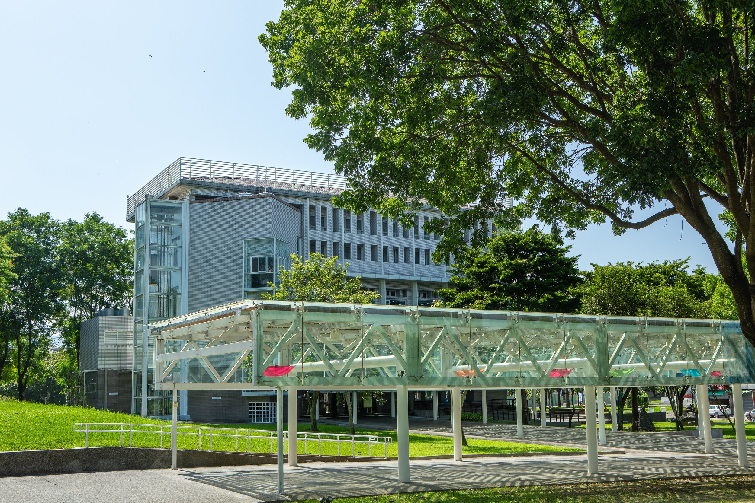 Modern building with glass elevators and a rooftop terrace, surrounded by green trees and grass on a sunny day.