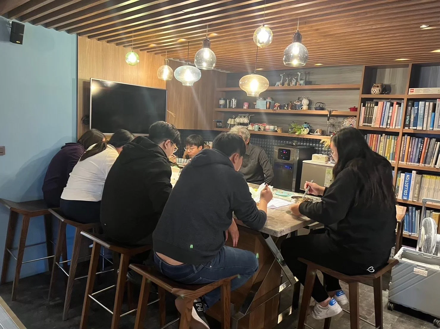 Group of people sitting at a wooden table, reading and working on papers, in a cozy room with bookshelves and hanging pendant lights.台中建築師事務所團隊日常聚餐與團隊合作氛圍