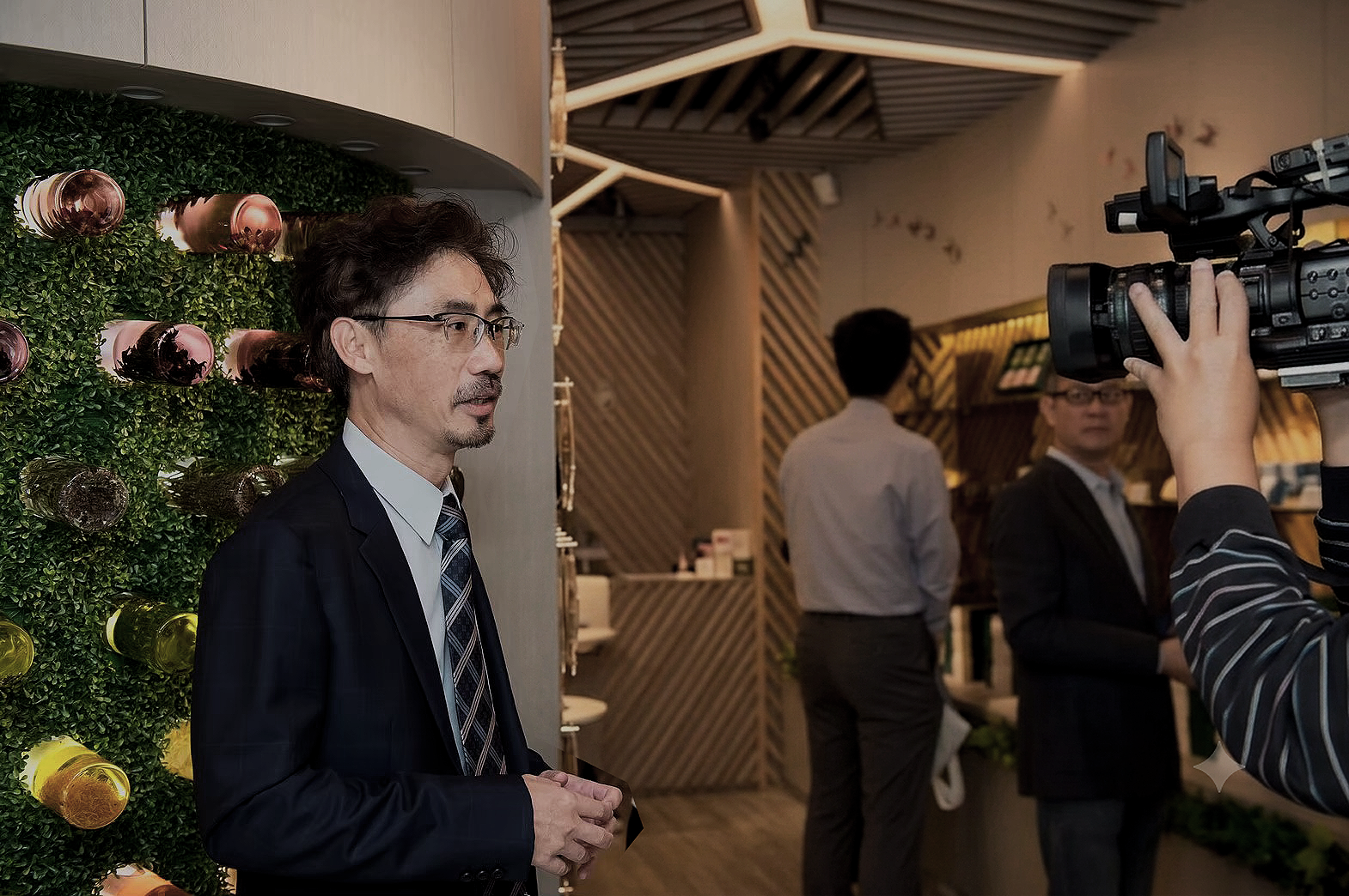 A man in a business suit standing in a modern interior, being filmed by a camera operator, with a wall of colorful decorative bottles behind him and other people in the background.