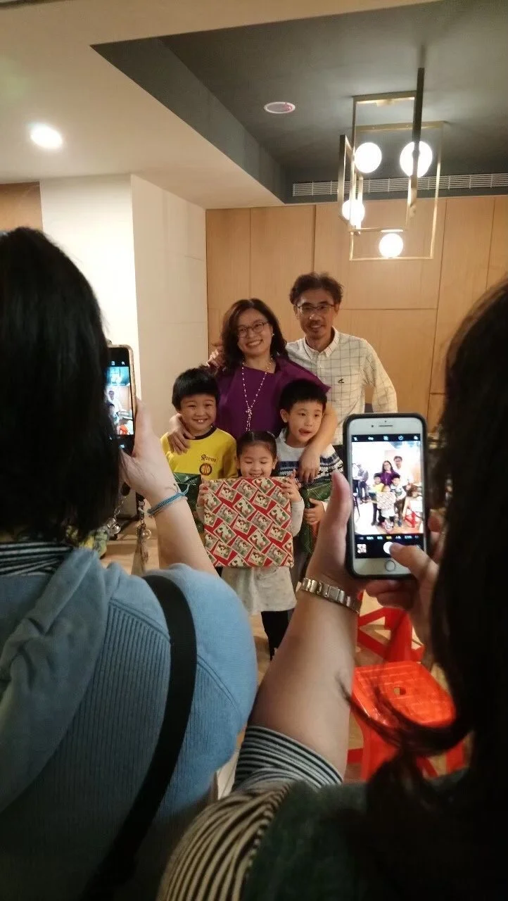 Group photo of two adults and four children at a celebration, with two women taking pictures on their smartphones in an indoor setting with wooden paneling and modern lighting.台中建築師事務所設計之溫馨木質感空間與客戶慶祝合影