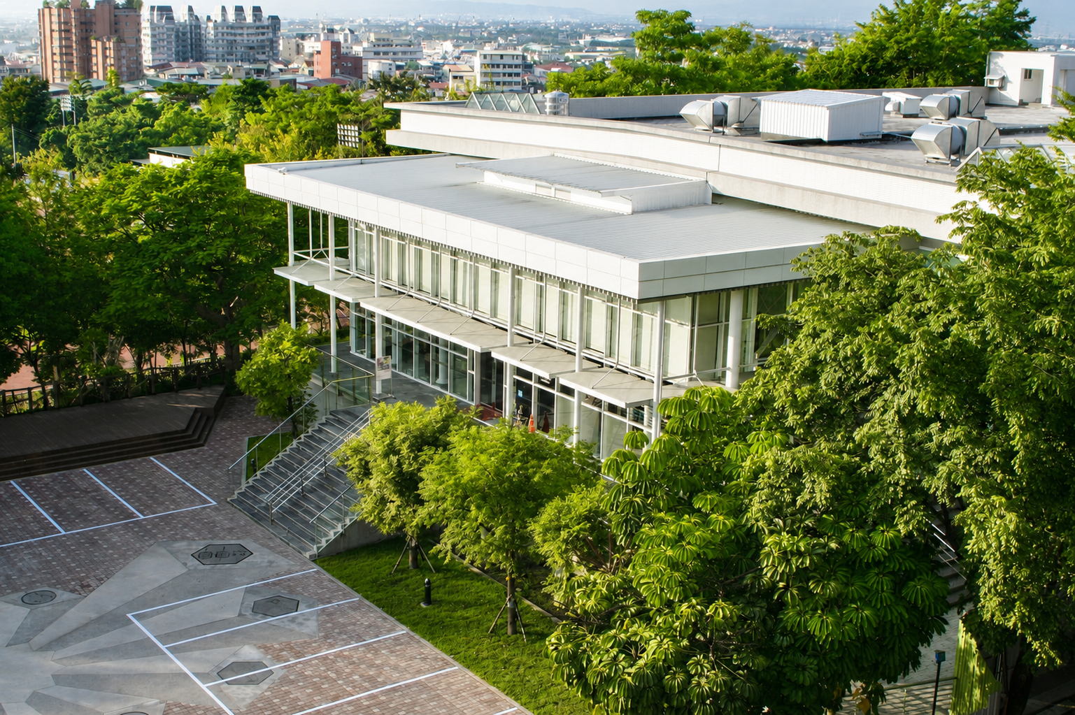 A modern building with large glass windows and a white roof, surrounded by lush green trees, with a parking area and cityscape in the background.