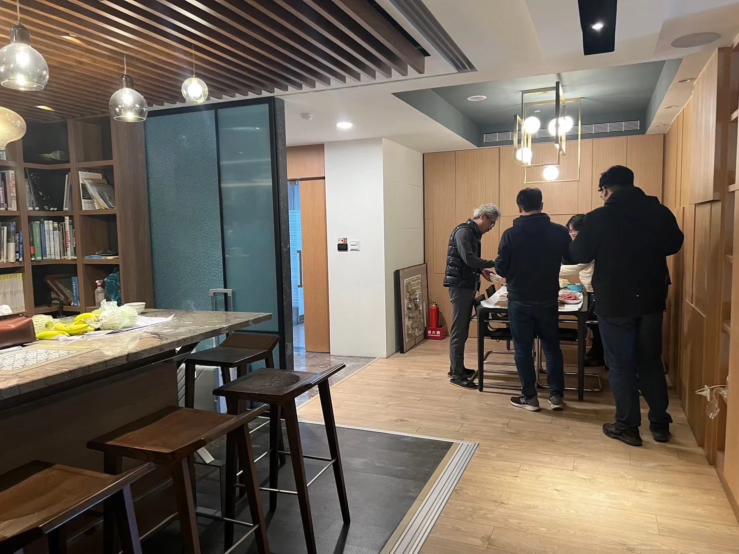 Group of five people standing around a table in a modern dining area, with wooden walls and hanging lights.台中建築師事務所團隊於現代木質會議區討論建築設計案