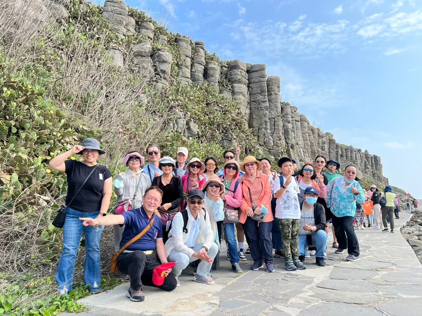 A large group of tourists posing for a photo on a stone pathway next to a rocky cliff under a partly cloudy blue sky. Many are wearing hats, sunglasses, and casual clothing.