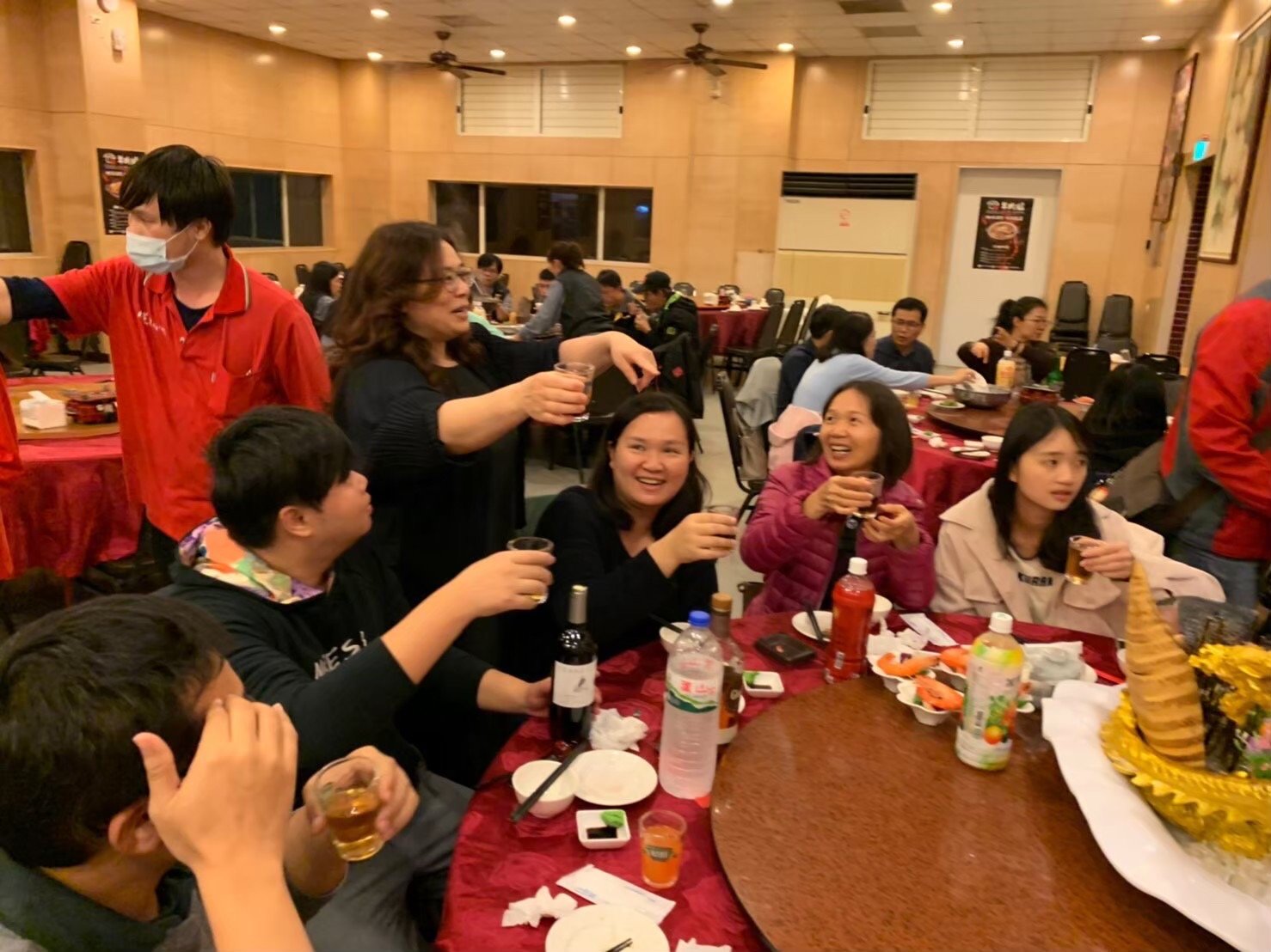 Group of people celebrating at a banquet, sitting around a table with drinks and food, some raising glasses in toast, in a decorated restaurant with red tablecloths and a large floral arrangement.台中建築師事務所團隊日常聚餐與團隊合作氛圍