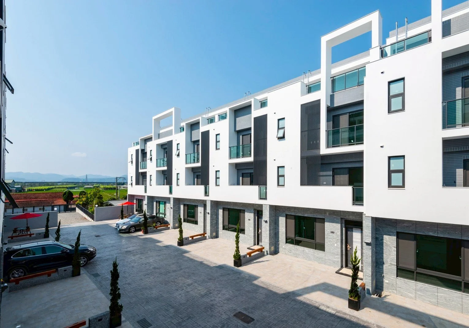 Modern white apartment building with balconies, large windows, and a parking lot with cars and benches outside.