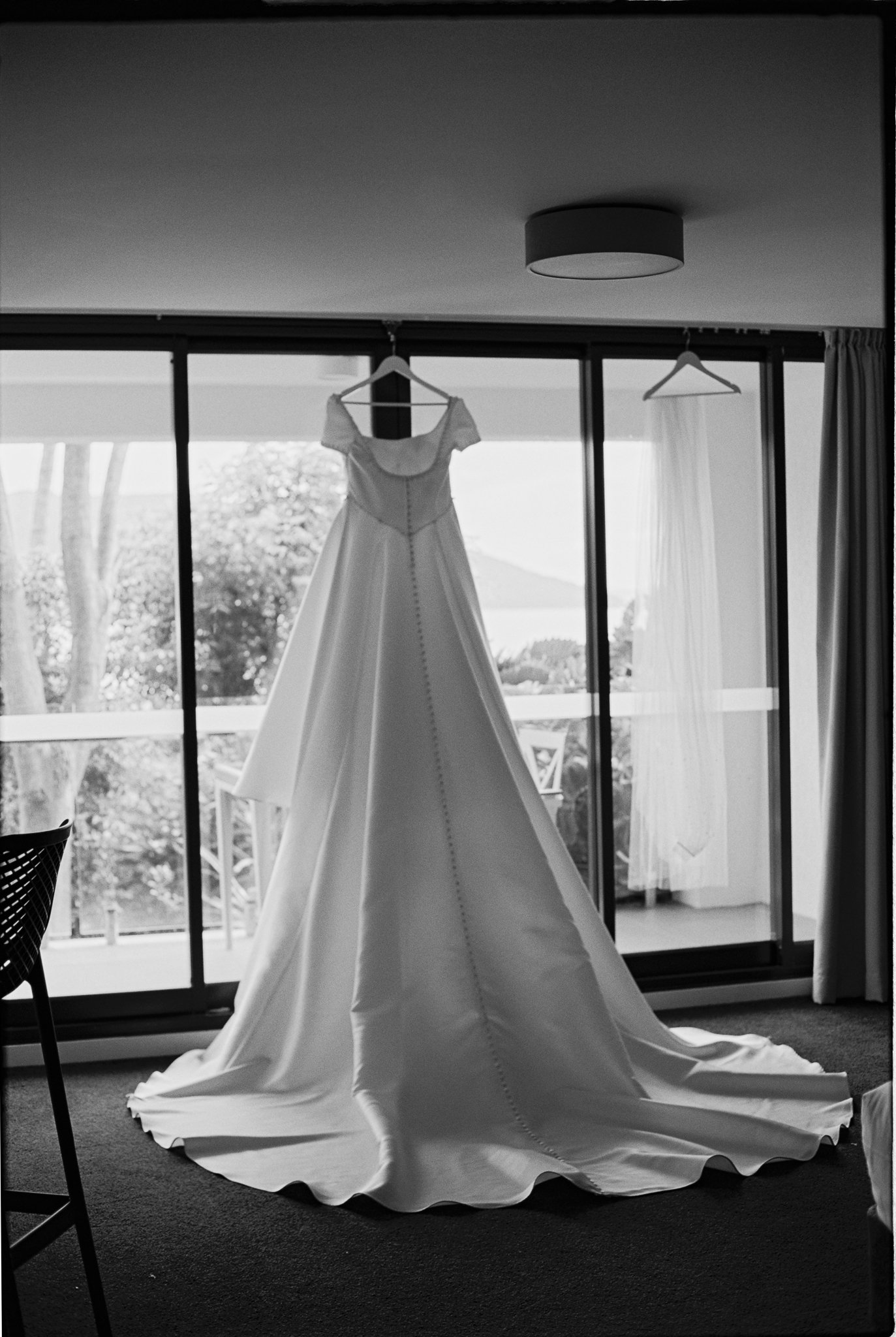 A wedding dress hanging on a hanger in front of sliding glass doors, with a landscape of trees and water visible outside. The dress is arranged with a long train on the floor.