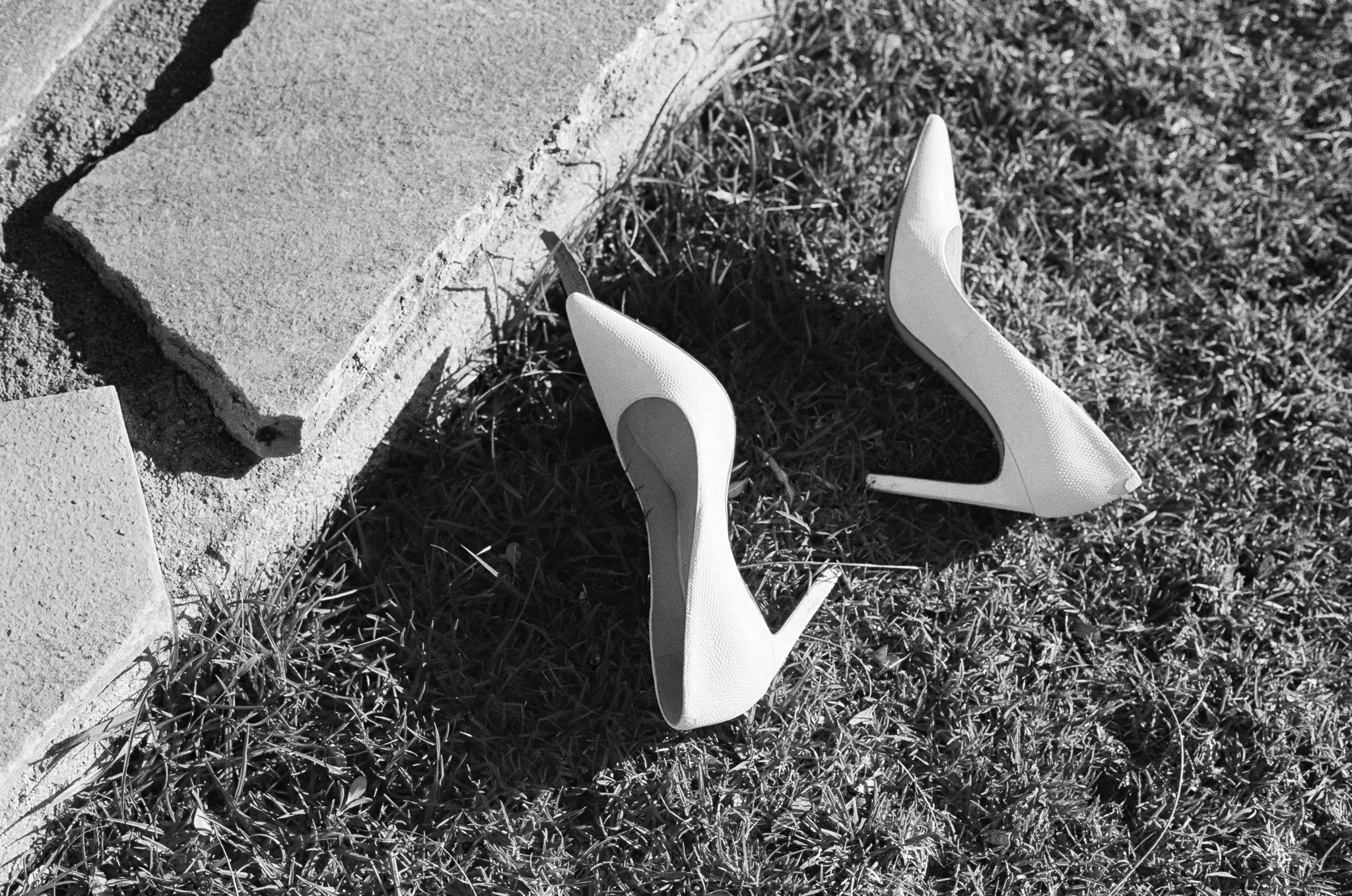 A pair of high-heeled shoes lying on grass near a concrete curb in black and white.