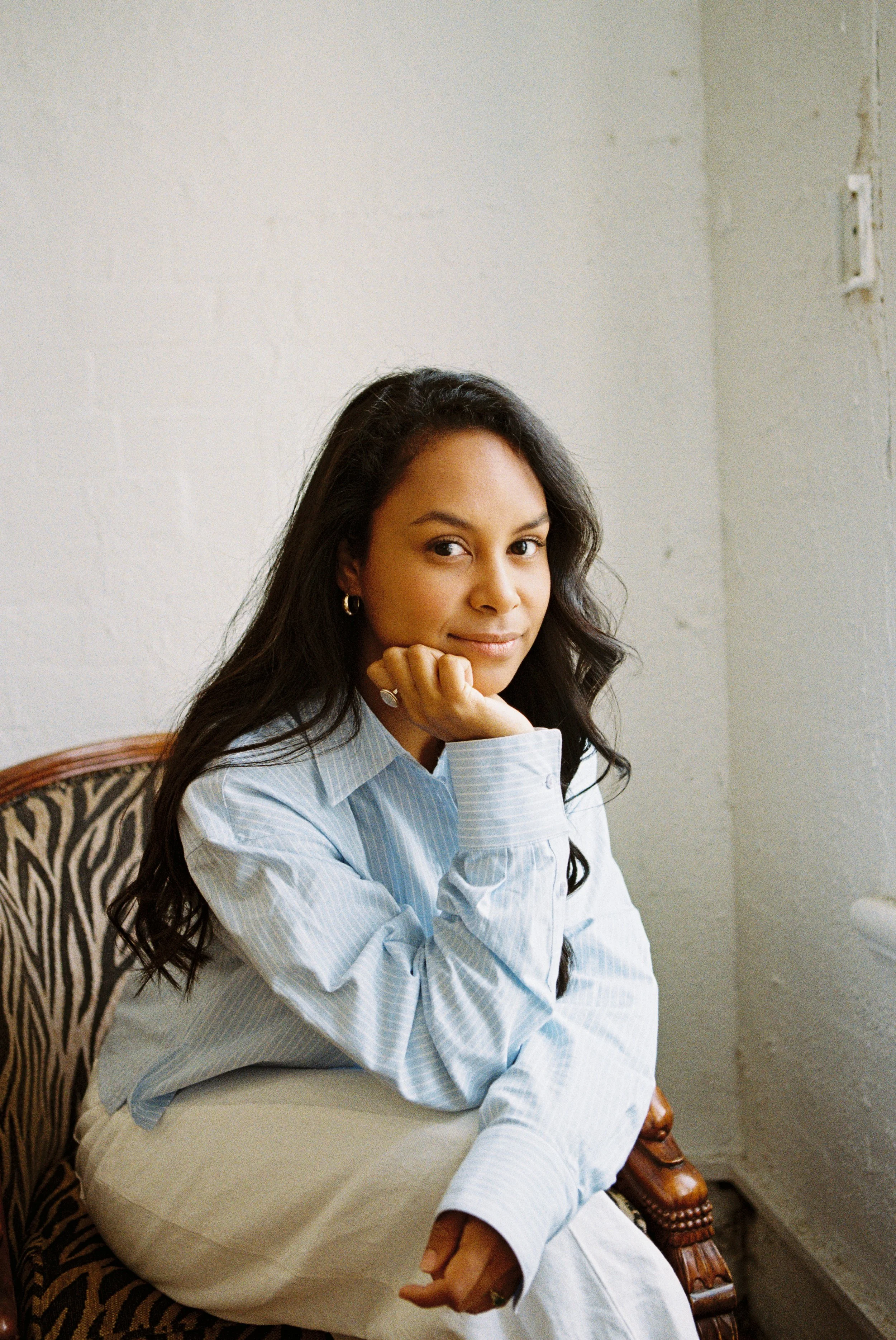 A young woman with long dark hair, wearing gold hoop earrings and a blue striped blouse, sitting on a patterned armchair. She rests her chin on her hand and looks slightly to the side with a subtle smile, in a room with a white brick wall and a grey wall with a small, open electrical outlet.