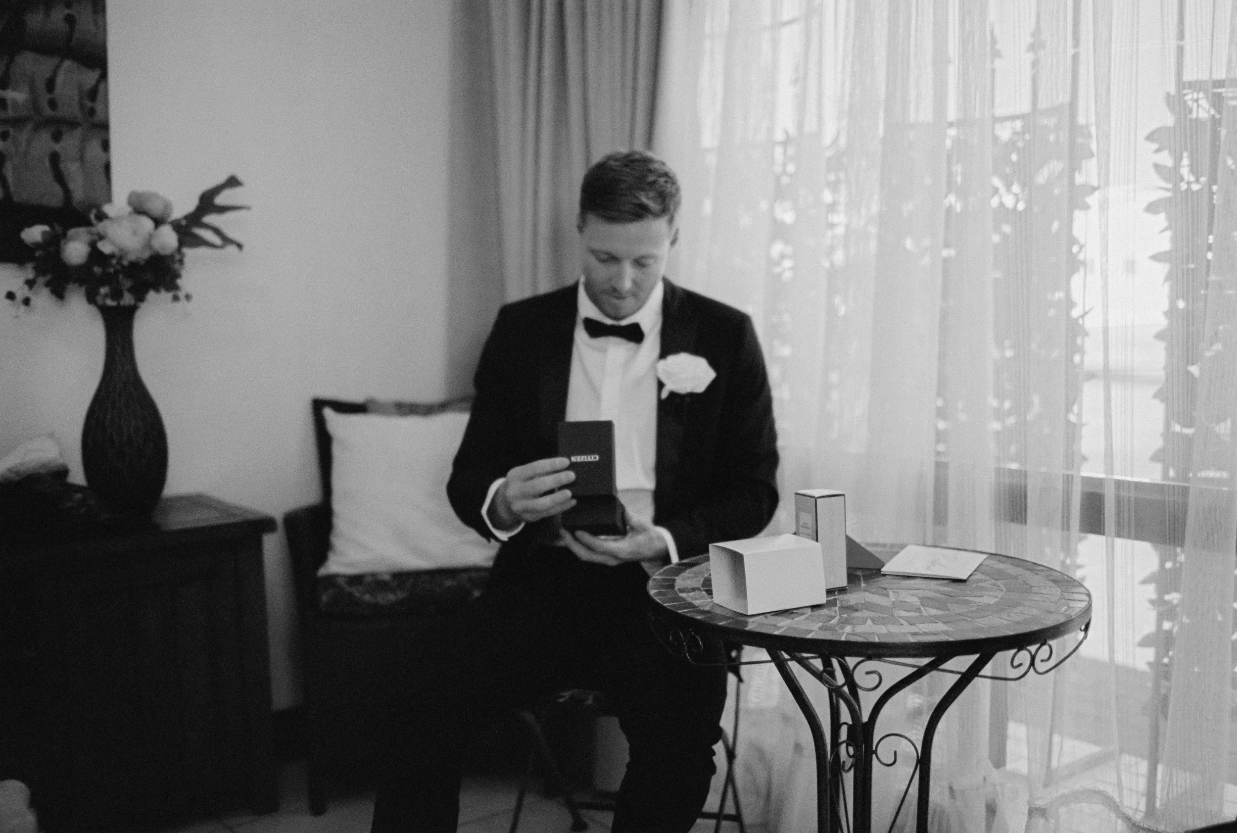 A young man dressed in a tuxedo with a bow tie and a white rose on his lapel is sitting at a round table, looking down at a small black box in his hands. There is a vase with flowers on a side table to the left and a window with curtains in the background.