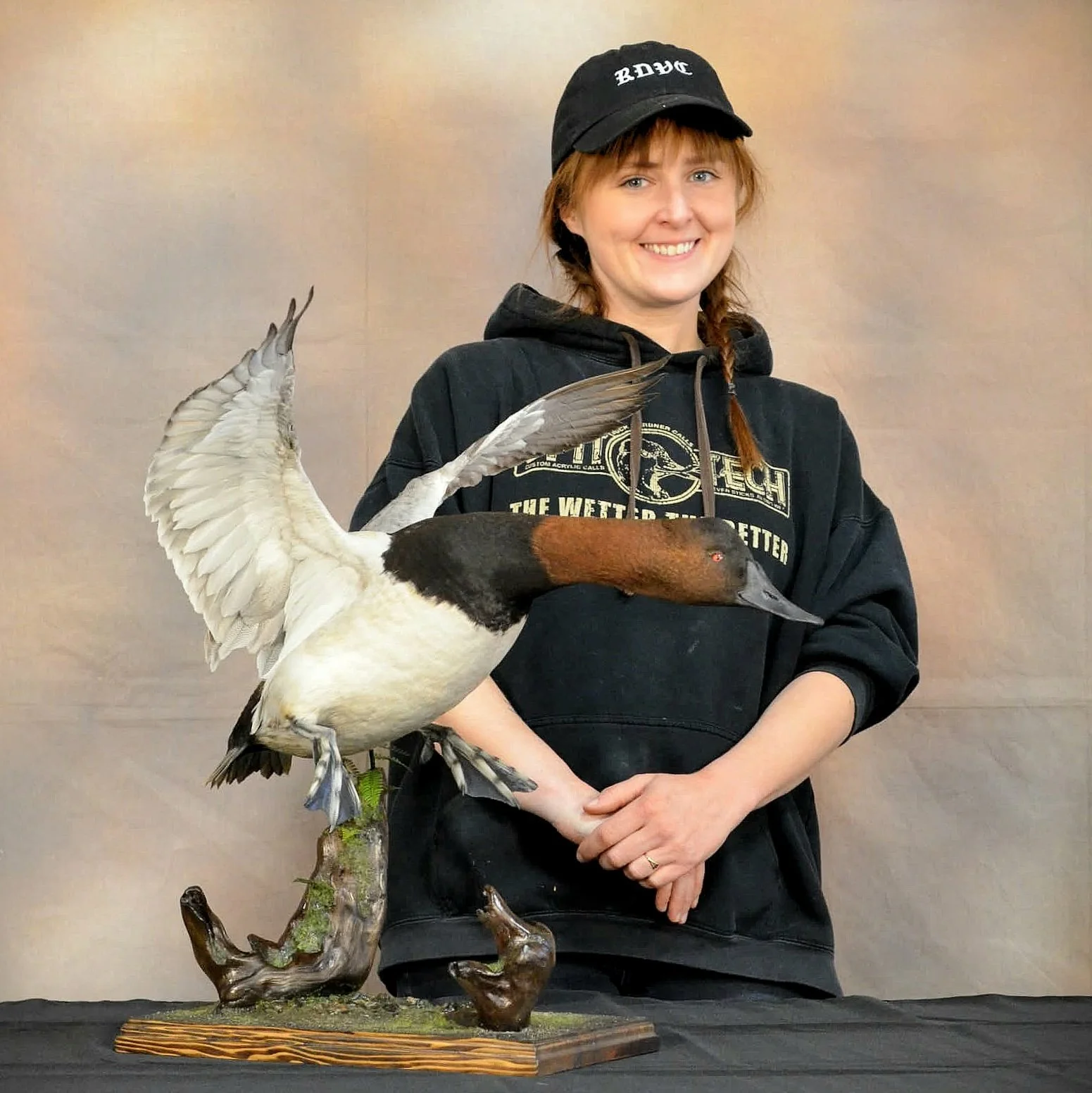 A young woman with red hair in braids wearing a black hoodie and black cap, smiling while holding a mounted duck and goose display on a wooden base.