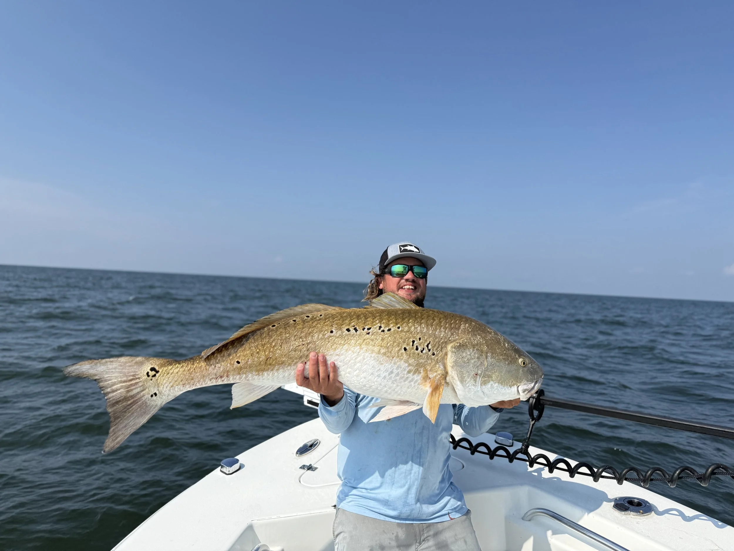 Man wearing sunglasses and a cap holding a large fish on a boat in the ocean under a clear blue sky.