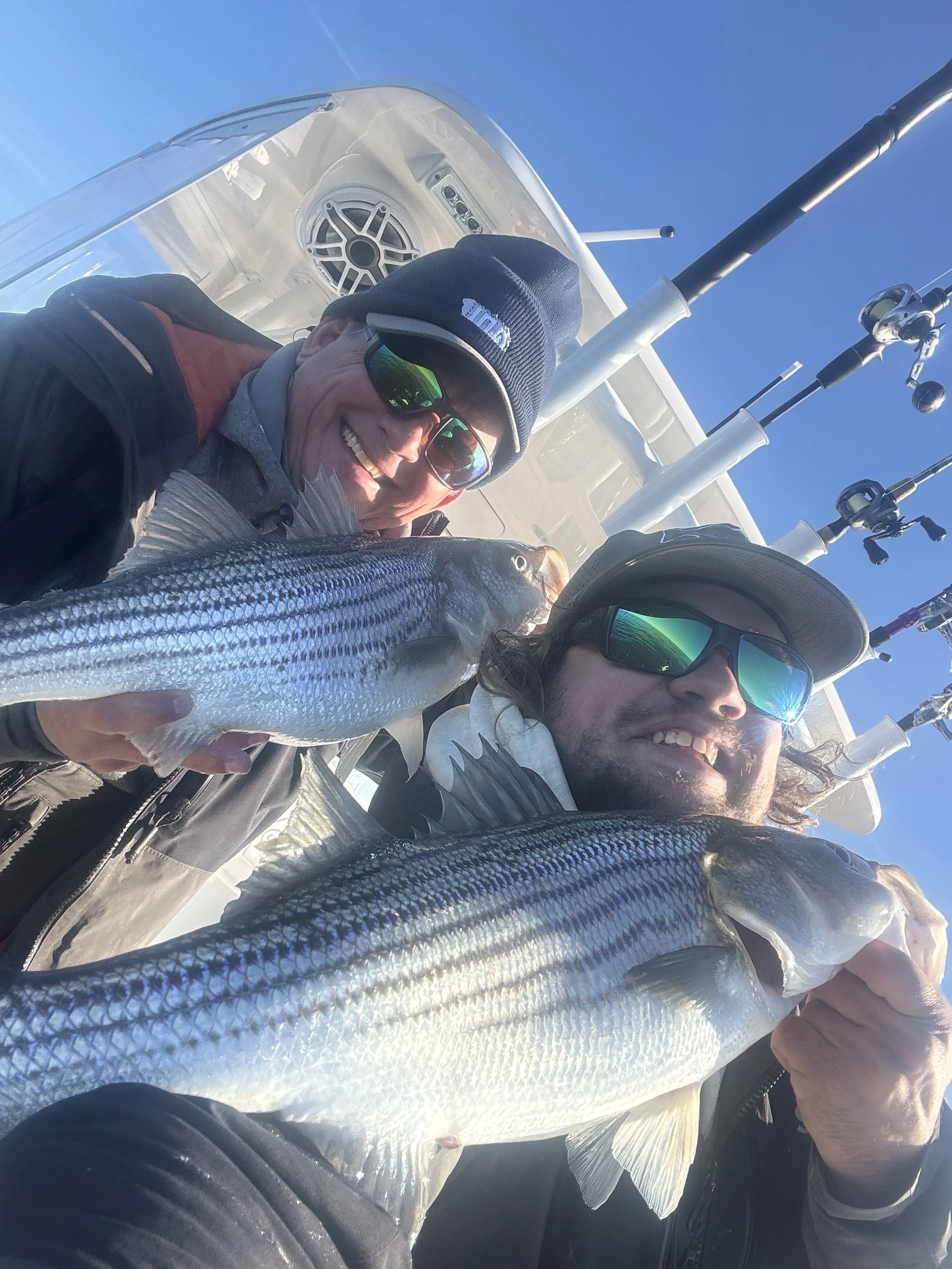 Two people on a boat holding a large fish, smiling, one wearing sunglasses and a navy beanie, the other with long hair and sunglasses, with fishing rods in the background.