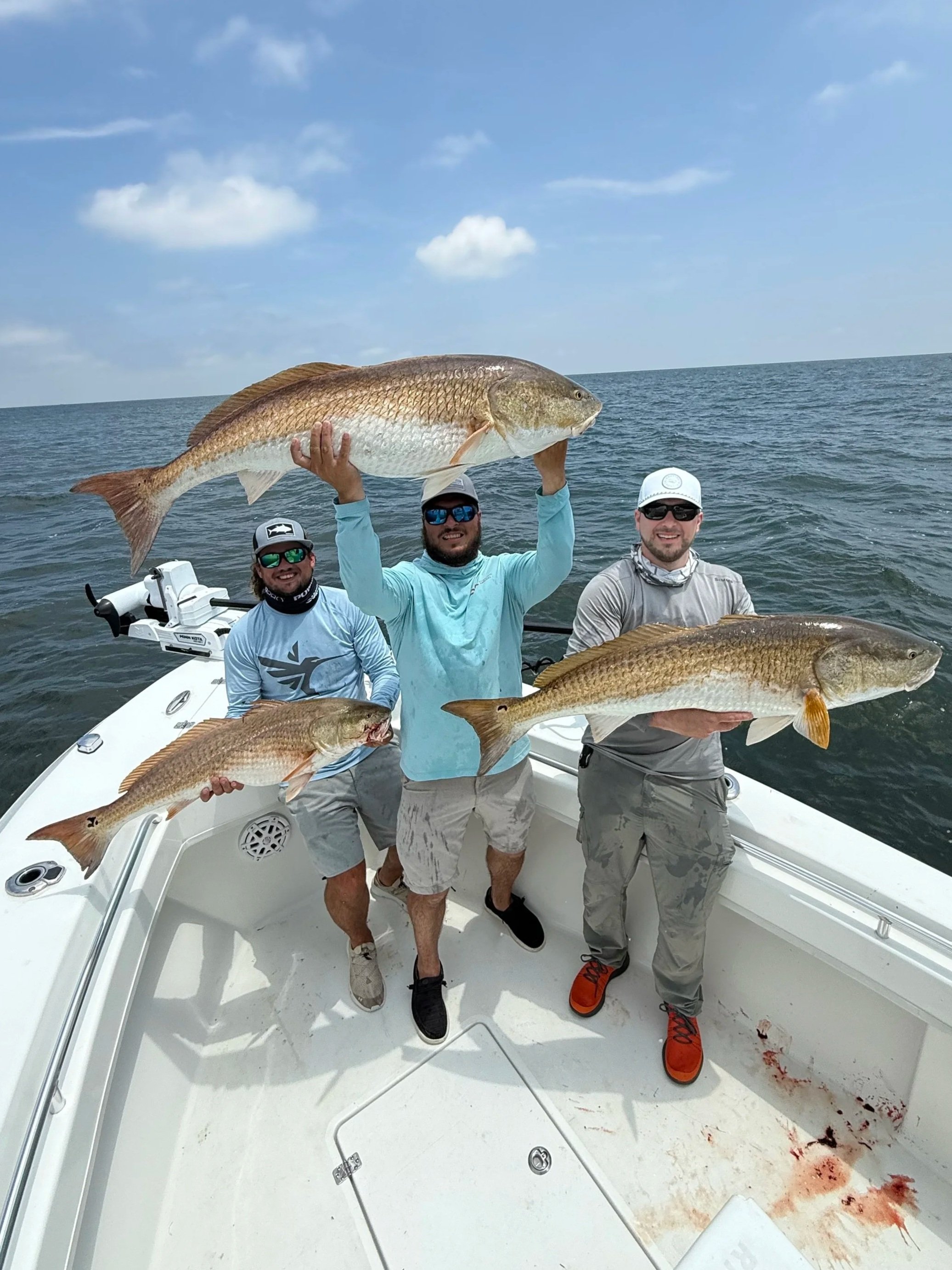 Three men on a boat holding large fish they caught. The boat is on open water, and there are clouds in the sky.