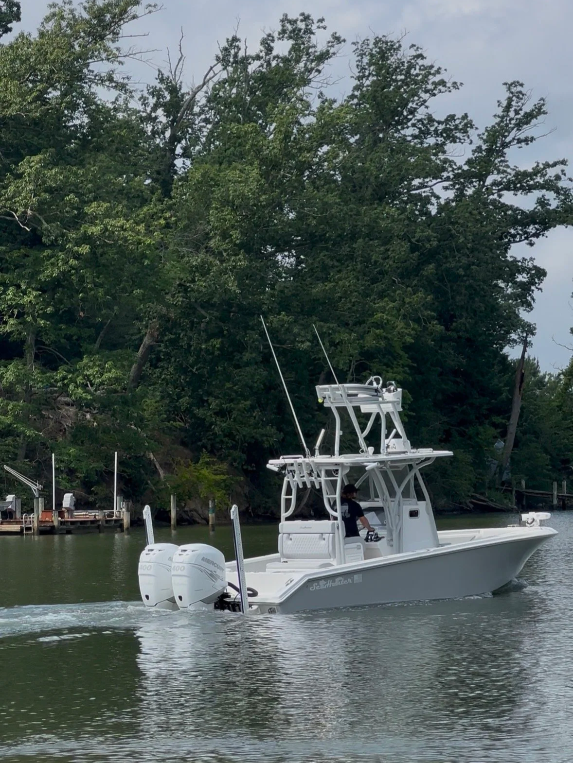 A white fishing boat with twin outboard motors moving across a lake with trees in the background.