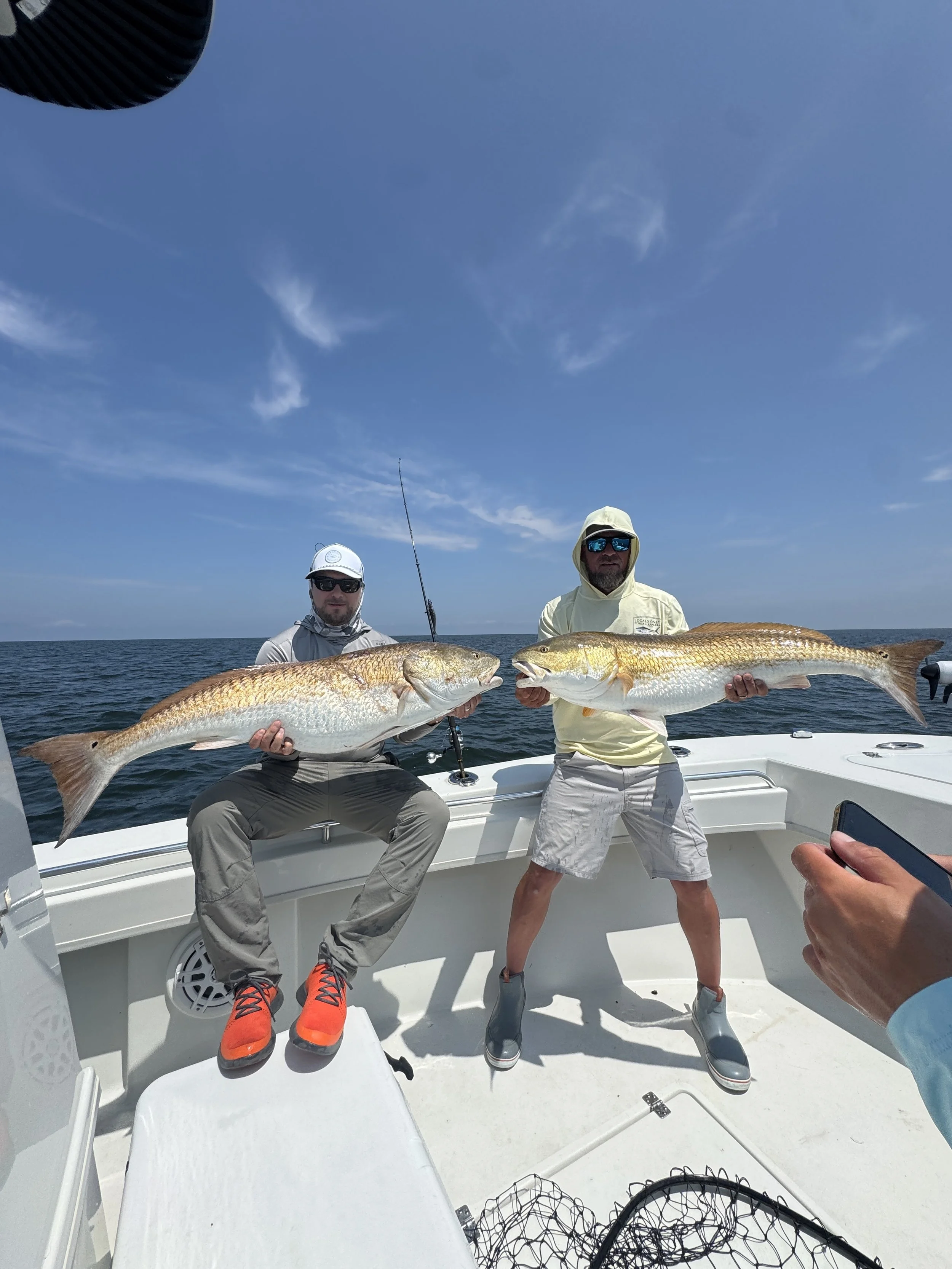 Two men on a boat holding large fish they caught, with blue sky and horizon in the background.