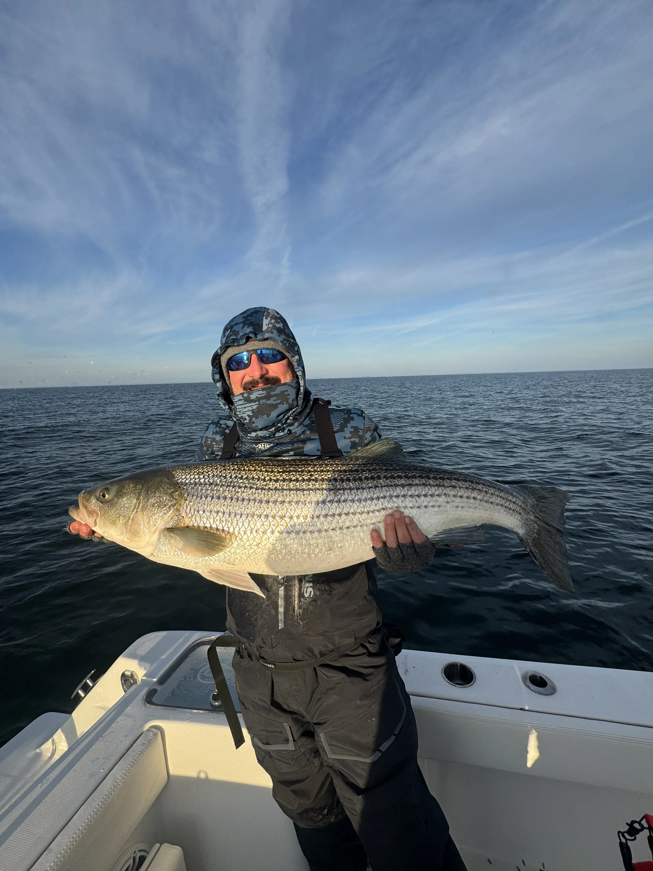 A man wearing camouflage jacket, dark pants, sunglasses, and a cap is standing on a boat holding a large fish. The background shows open water and a blue sky with thin clouds.