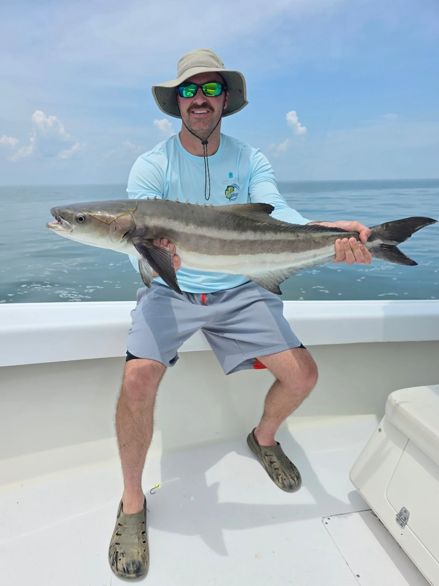 A man wearing a wide-brimmed hat and sunglasses holding a large fish on a boat, with the ocean and sky in the background.
