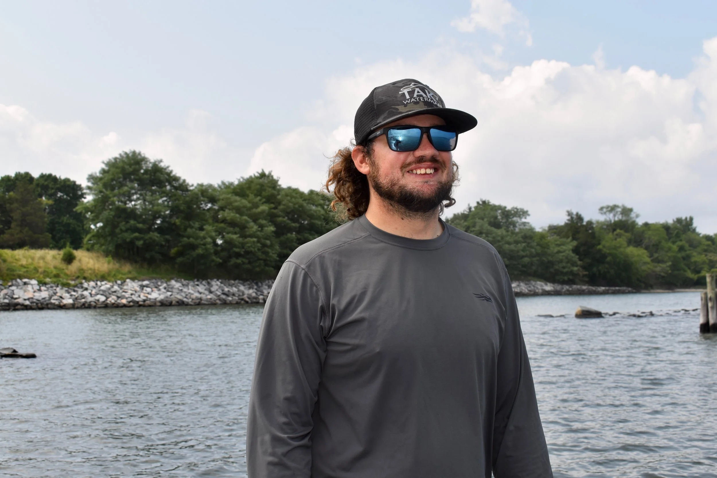 A man with curly hair and a beard, wearing sunglasses, a camouflage cap, and a gray long-sleeve shirt, standing outdoors near a body of water with trees in the background.