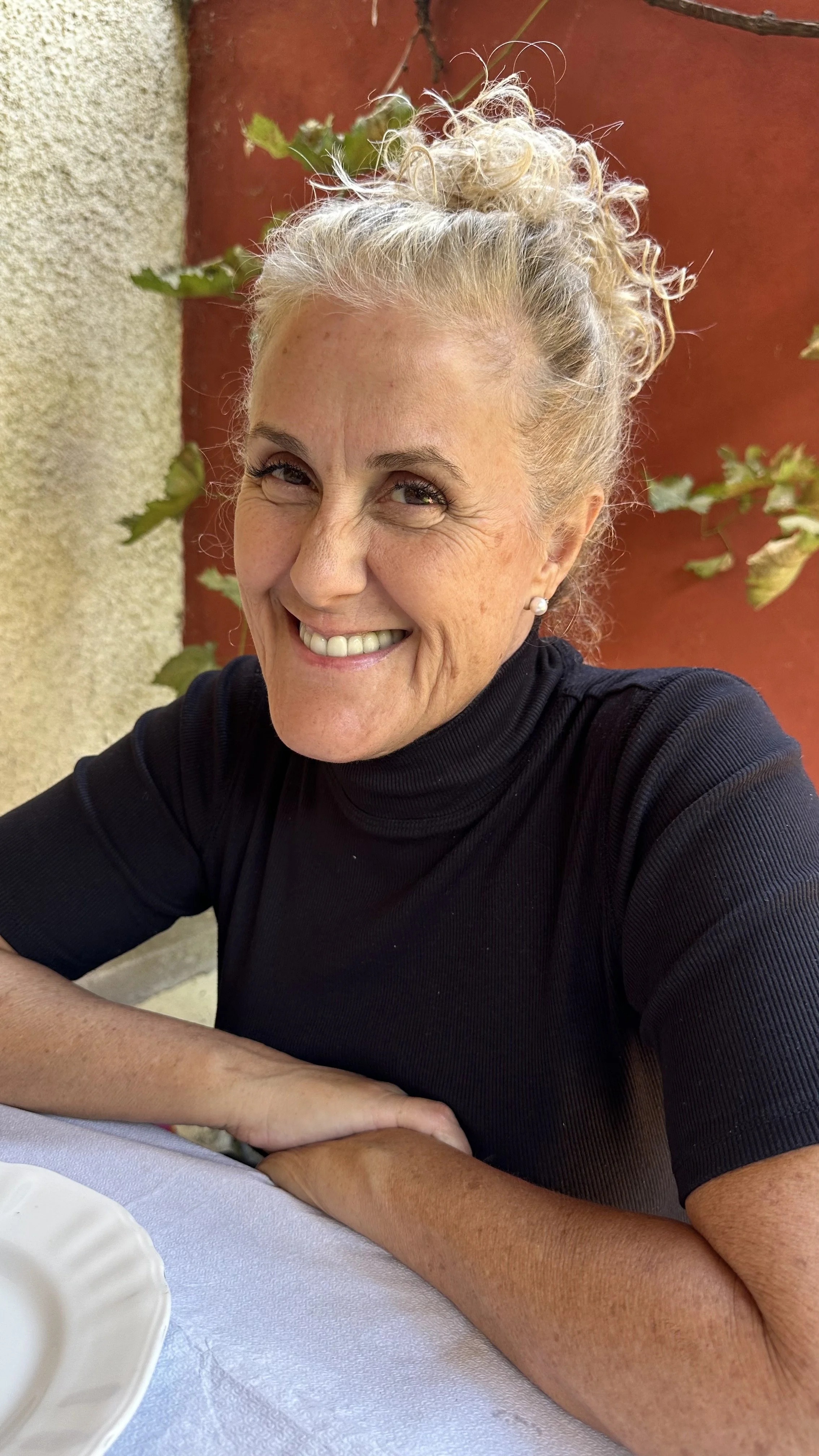 A smiling woman with curly blonde hair styled in an updo, wearing a black turtleneck and pearl earrings, sitting at a table outdoors with a red wall and green leaves in the background.