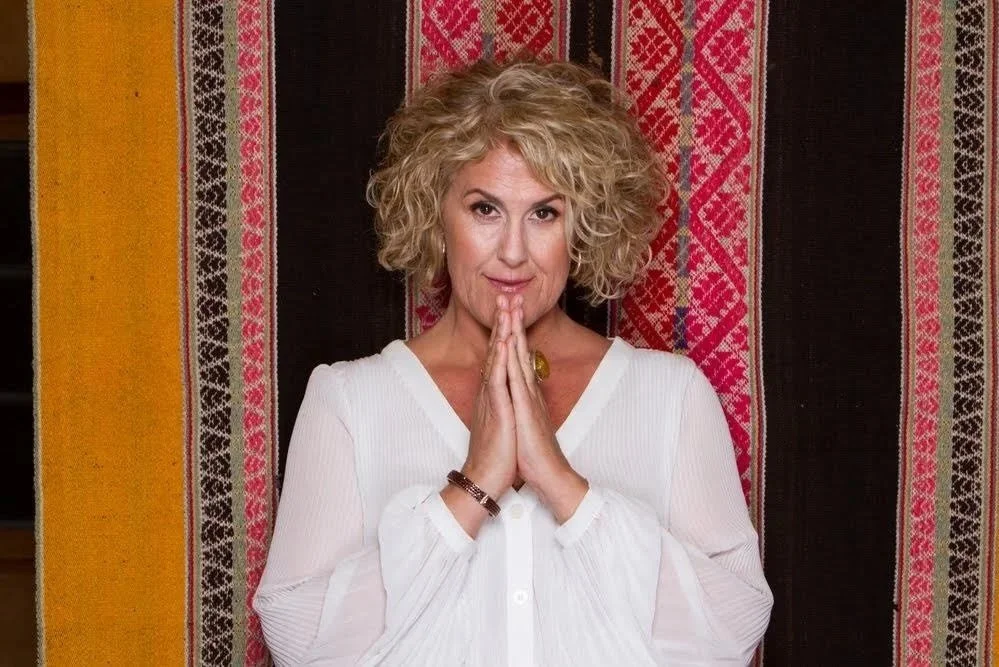 A woman with blonde curly hair, wearing a white blouse, standing in front of a colorful woven textile background, with her hands pressed together in a gesture of prayer or gratitude.