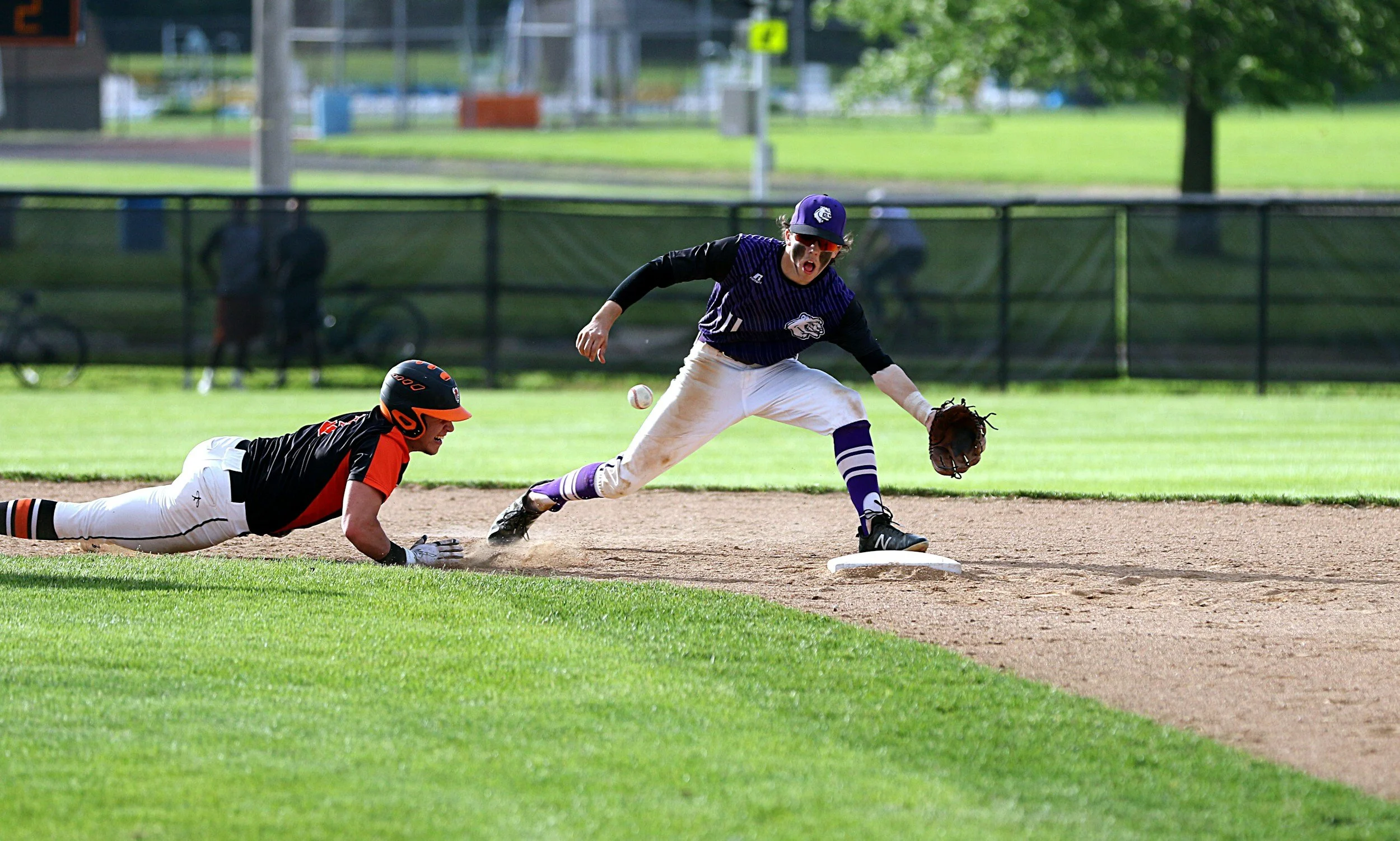 A baseball player in black and orange attempting to reach a base while a player in purple and white makes a play to tag him out during a game on the field.