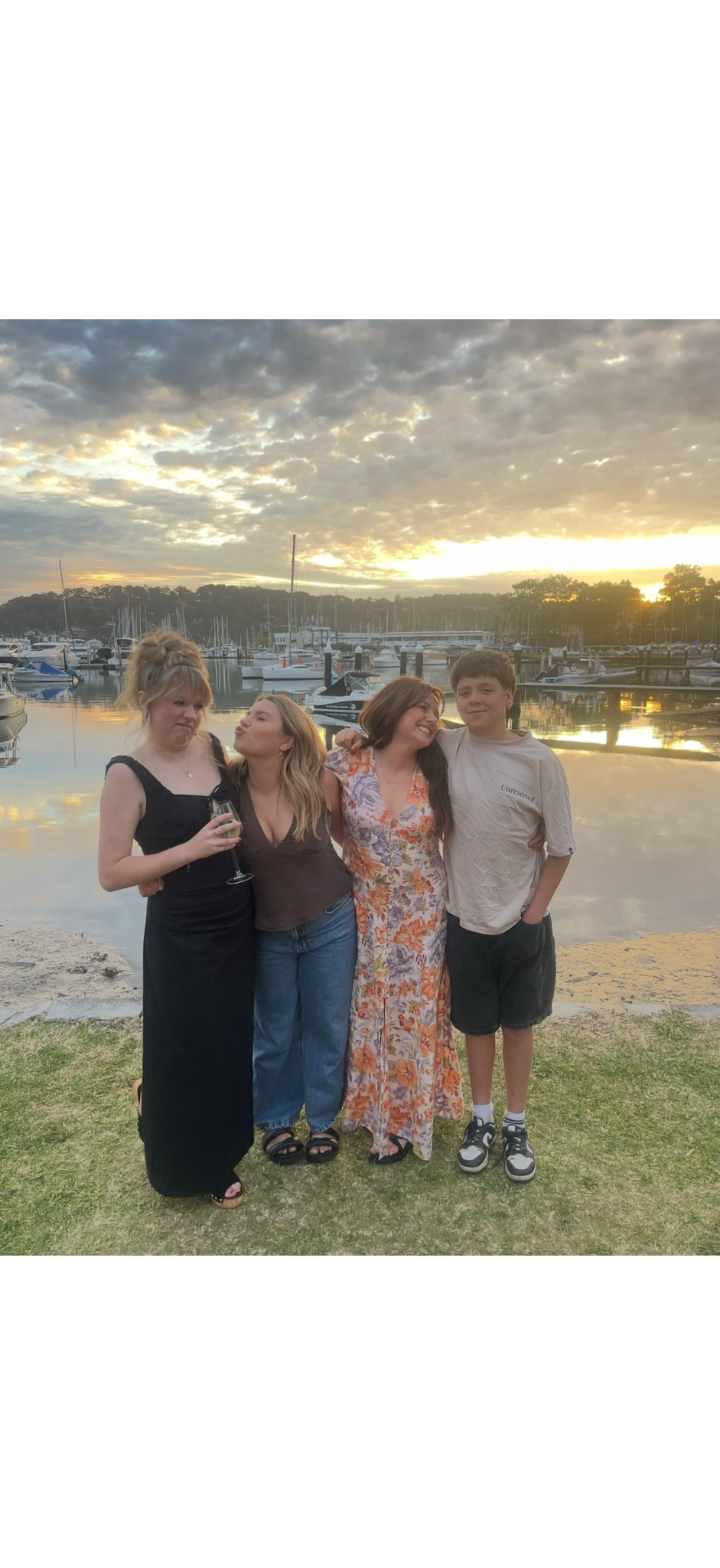Four people standing together by a marina with boats and a sunset sky in the background, smiling and making funny faces.