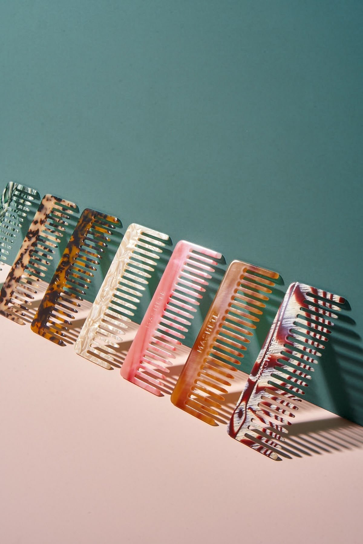 Six colorful hair combs with various marbled and solid patterns, lined up diagonally on a white surface, with shadows cast on a green and white background.