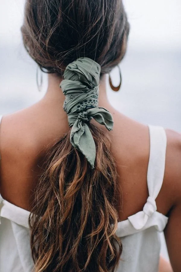 Back of a woman with wavy brown hair tied in a ponytail with a green patterned scarf, wearing hoop earrings and a white sleeveless top.