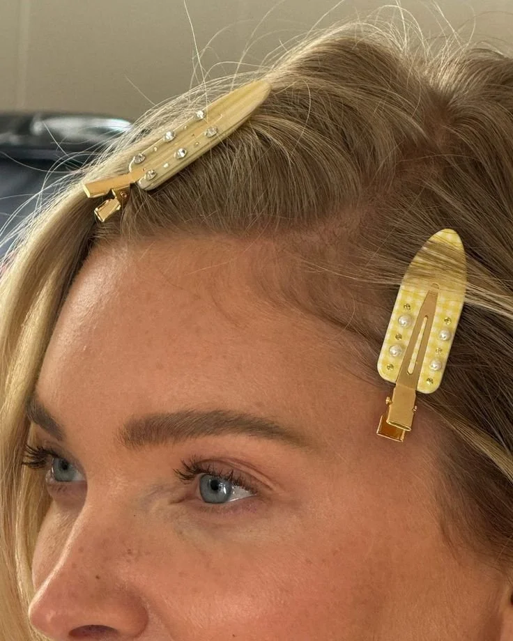Close-up of a woman's forehead with two yellow hair clips decorated with pearls and rhinestones in her blonde hair.