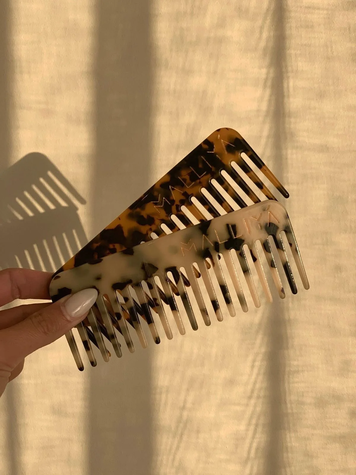 Two tortoiseshell patterned hair combs with one being darker and the other lighter, held by a hand with painted nails, casting shadows on a light background.
