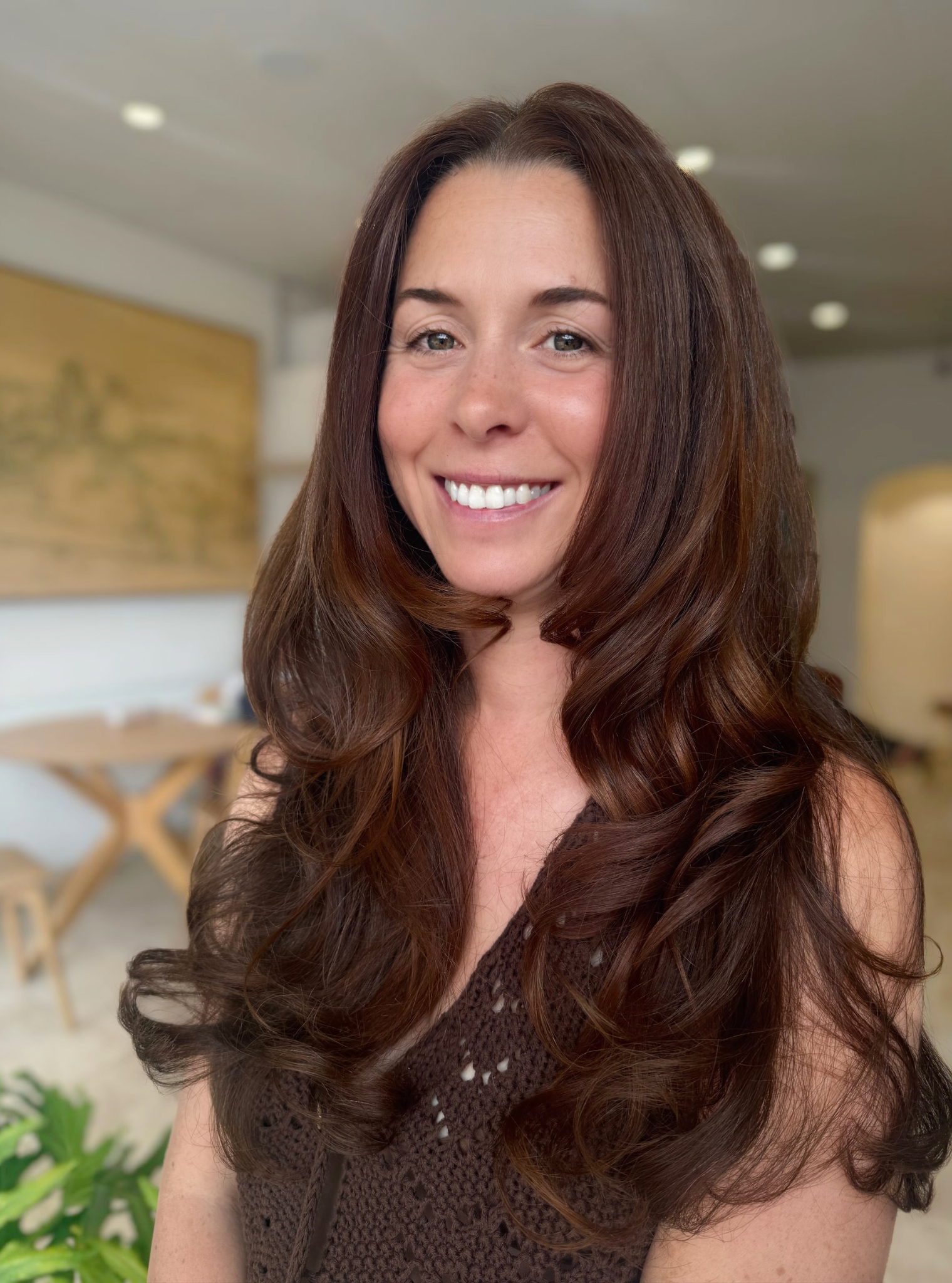 Close-up of a smiling woman with long, wavy brown hair, wearing a brown crochet top, indoors with blurred background.