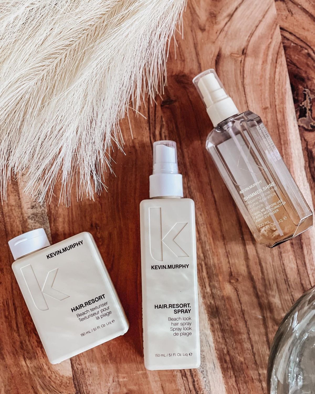 Three Kevin.Murphy hair products on a wooden surface: a bottle of HAIR.RESORT. Beach attend hue, a spray bottle of HAIR.RESORT. Spray, and a smaller bottle of HAIR.RESORT. Beach attend hue. There is also a white feather nearby.