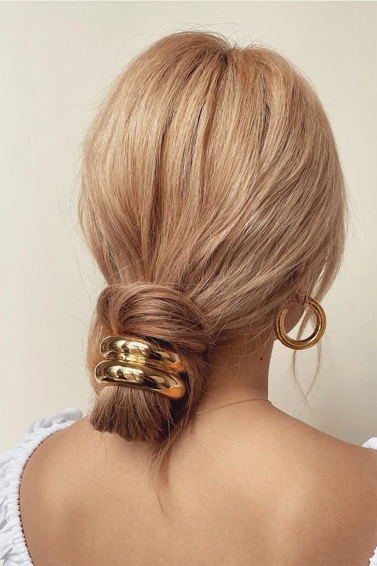 Back view of a woman's blonde hair styled in a low bun with large gold hair clips. She is wearing gold hoop earrings and a white top.