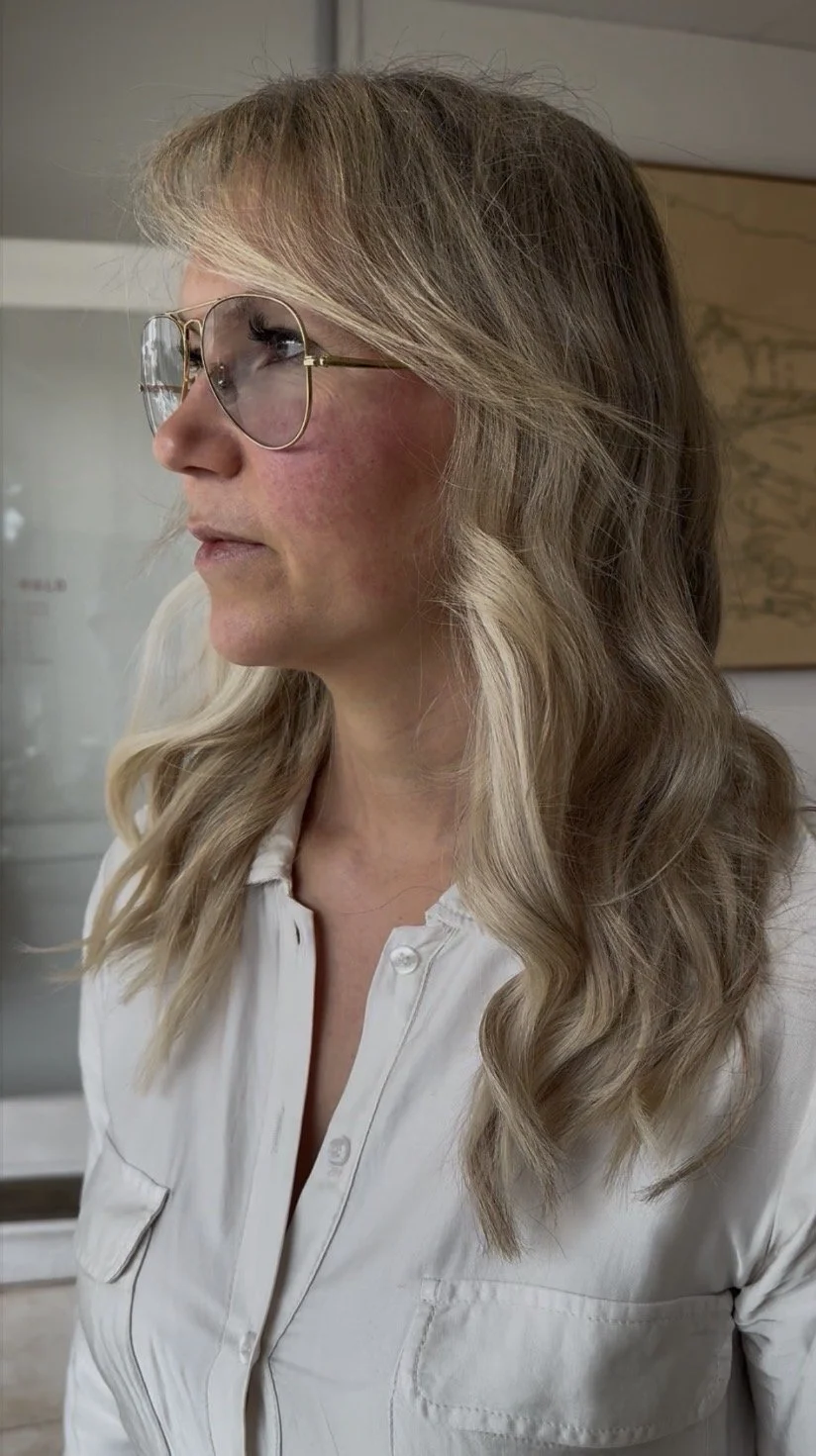Side view of a woman with blonde wavy hair wearing sunglasses and a white button-up shirt.