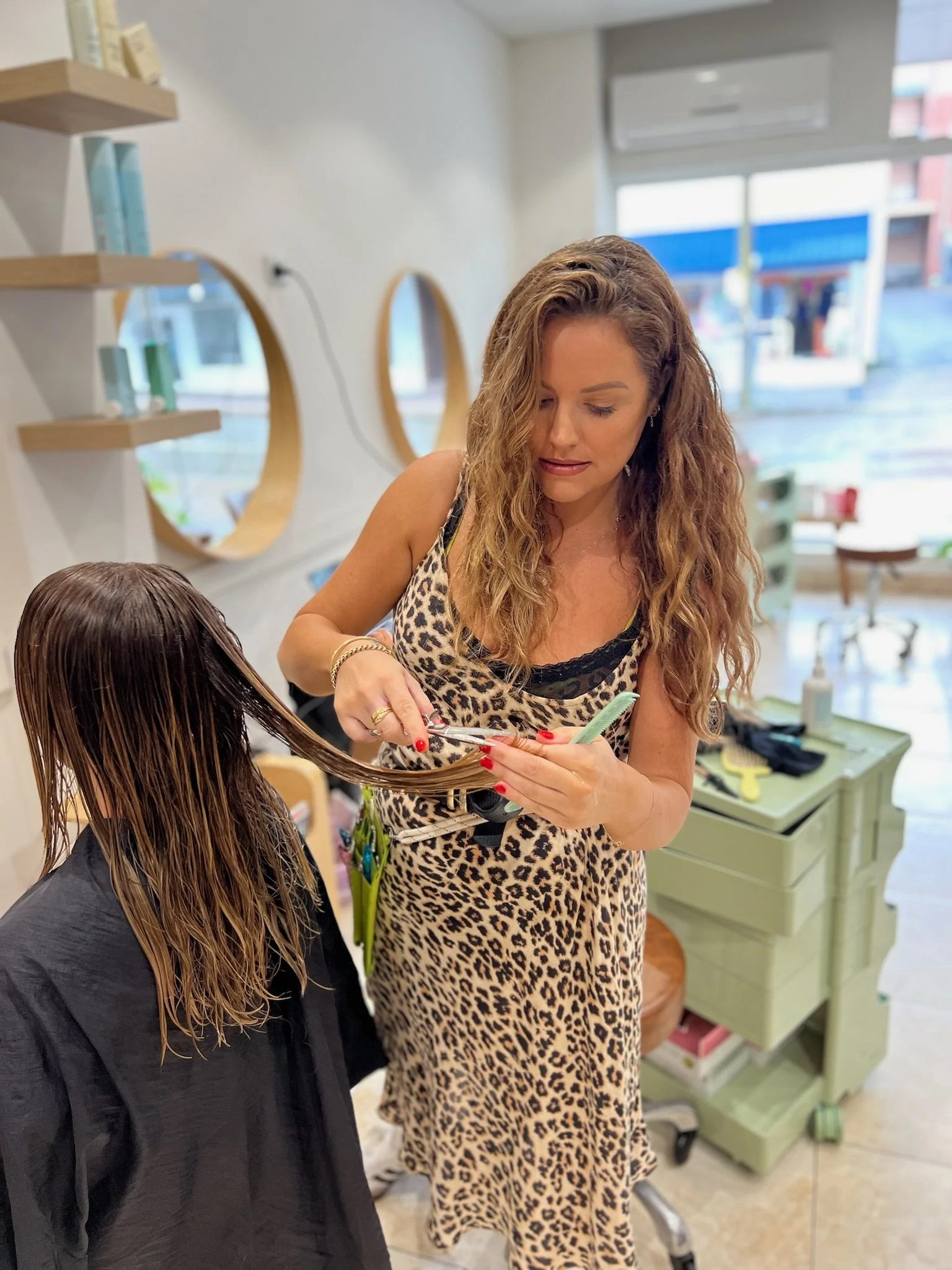 A woman with long curly hair and a leopard print dress cutting the hair of a young girl with long dark hair at a salon. The salon has a mirror and shelves with hair products, and a window showing buildings outside.