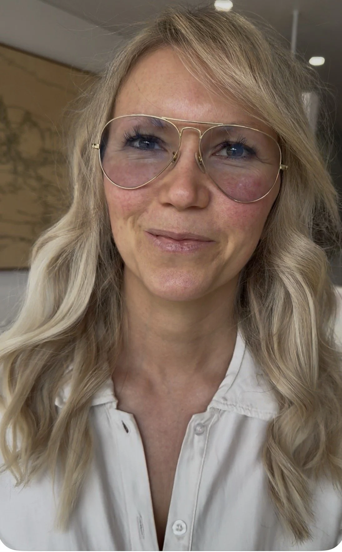 Close-up of a woman with long, wavy blonde hair wearing large aviator glasses and a white shirt, smiling softly.