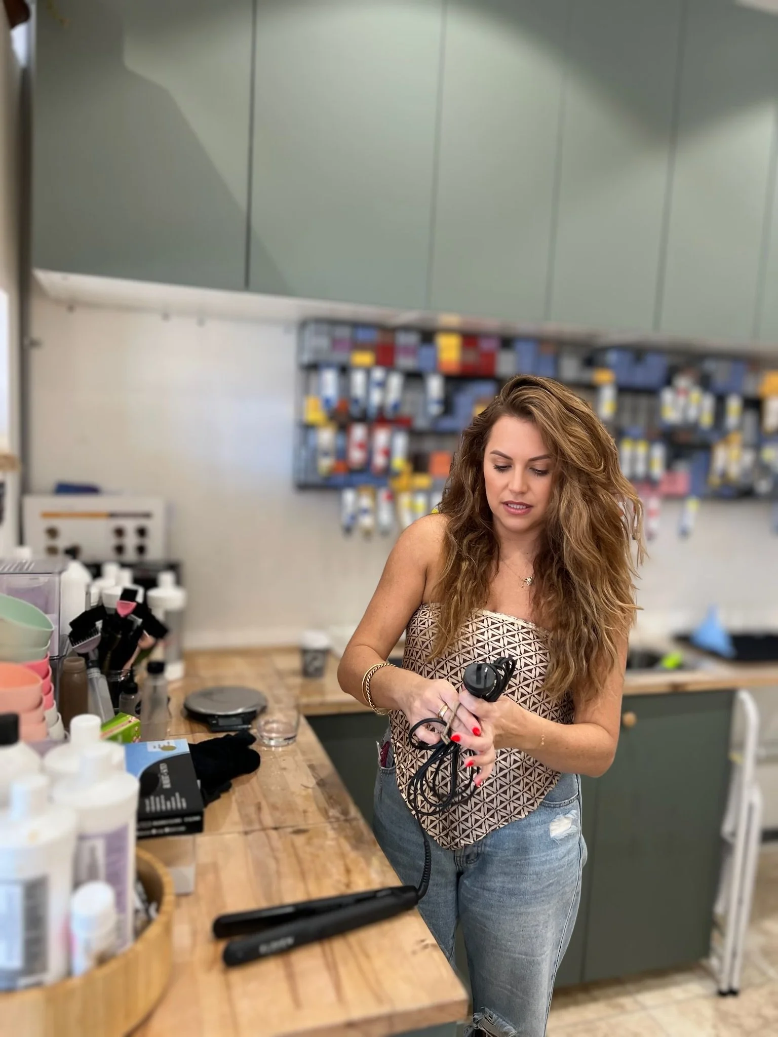 A woman with long curly hair, wearing a patterned sleeveless top and blue jeans, is holding a black electric device in a pharmacy or retail store with shelves of products and appliances in the background.