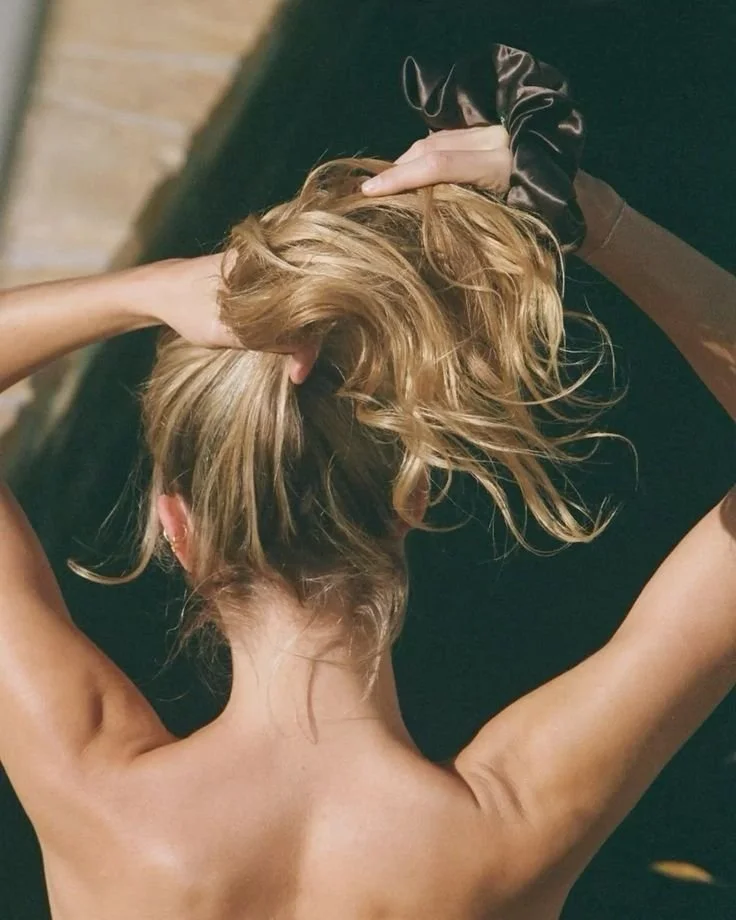 A woman with blonde hair styled in a messy bun, seen from behind, with her arms raised and hands resting on her head.