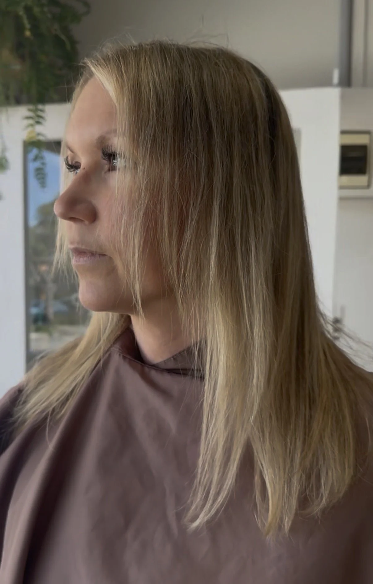 A woman with long, straight blonde hair is sitting in a salon, looking to the left, wearing a brown cape.