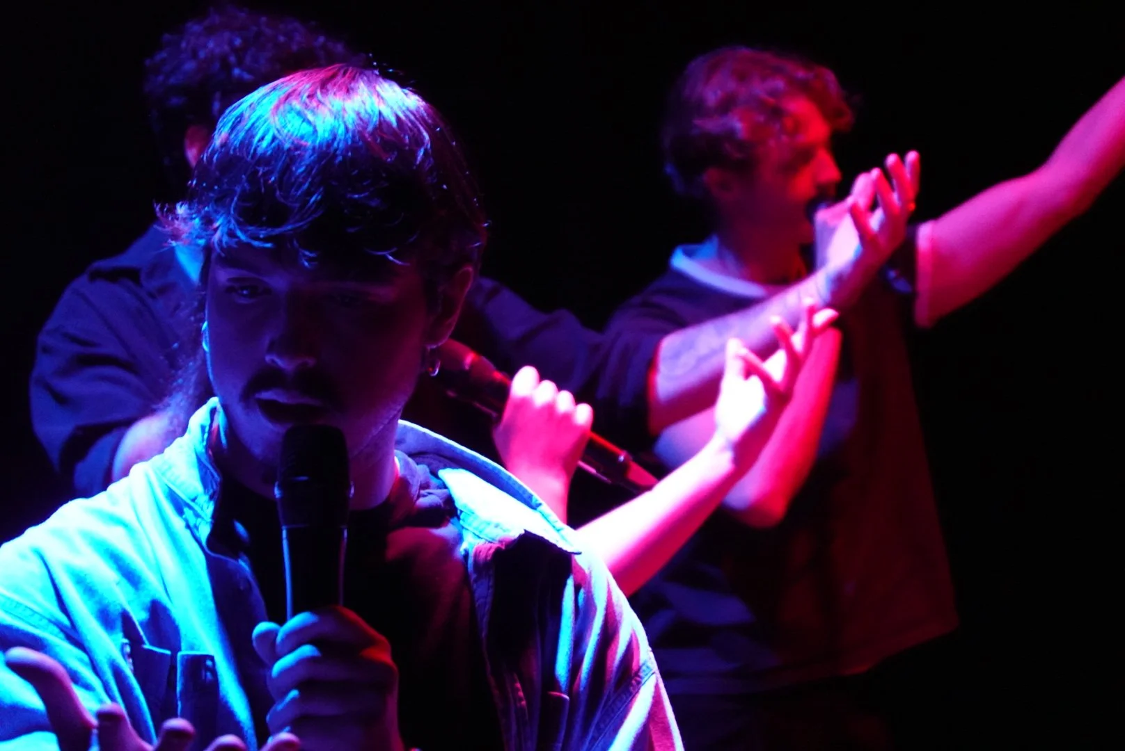 Two young men on stage, one in the foreground holding a microphone and the other in the background holding a microphone and gesturing with his hand, under colorful stage lighting.