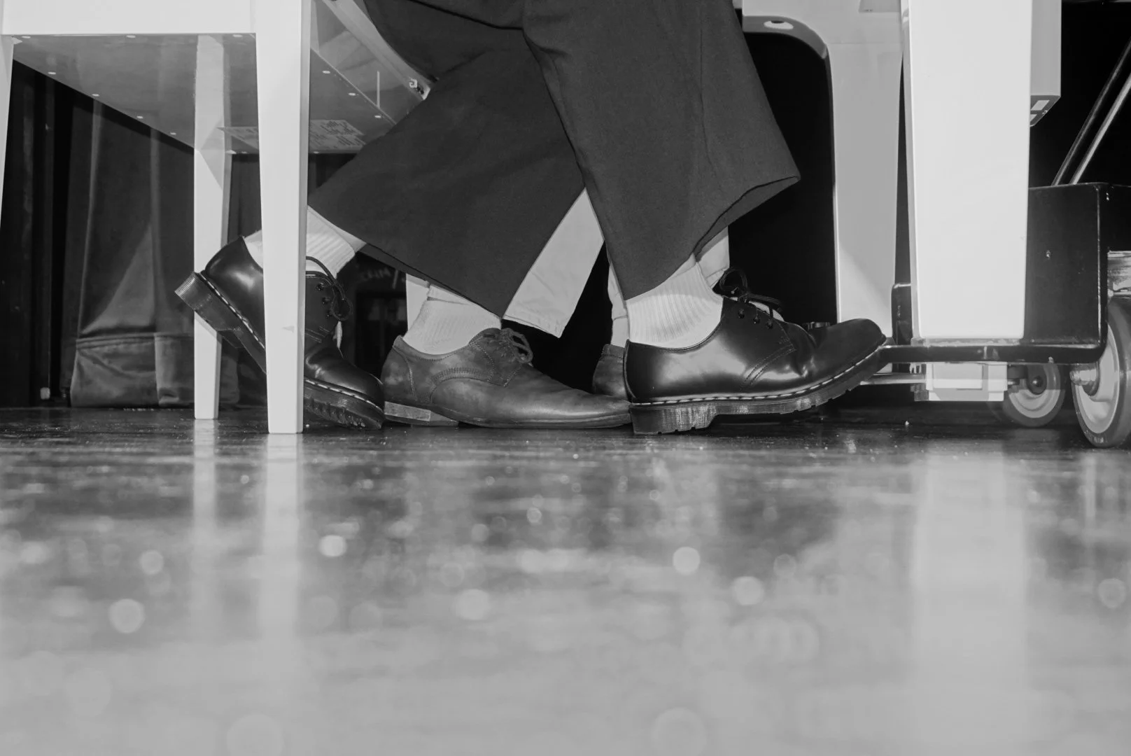 Close-up of two people's shoes under a table; one person is wearing polished black dress shoes with white socks, the other is wearing worn brown shoes with black pants. The setting appears to be indoors.