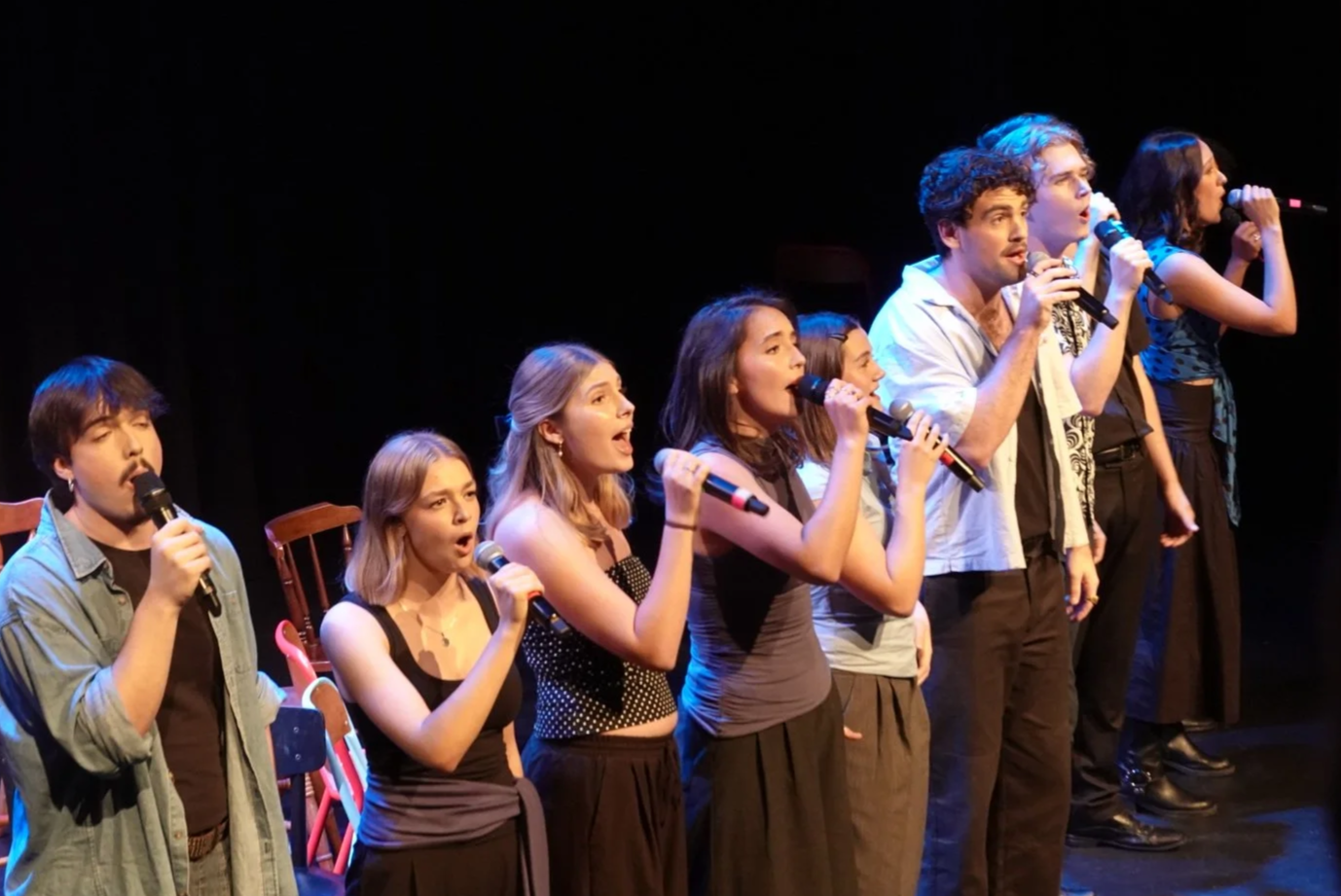 Group of diverse people singing into microphones on stage during a performance or rehearsal.