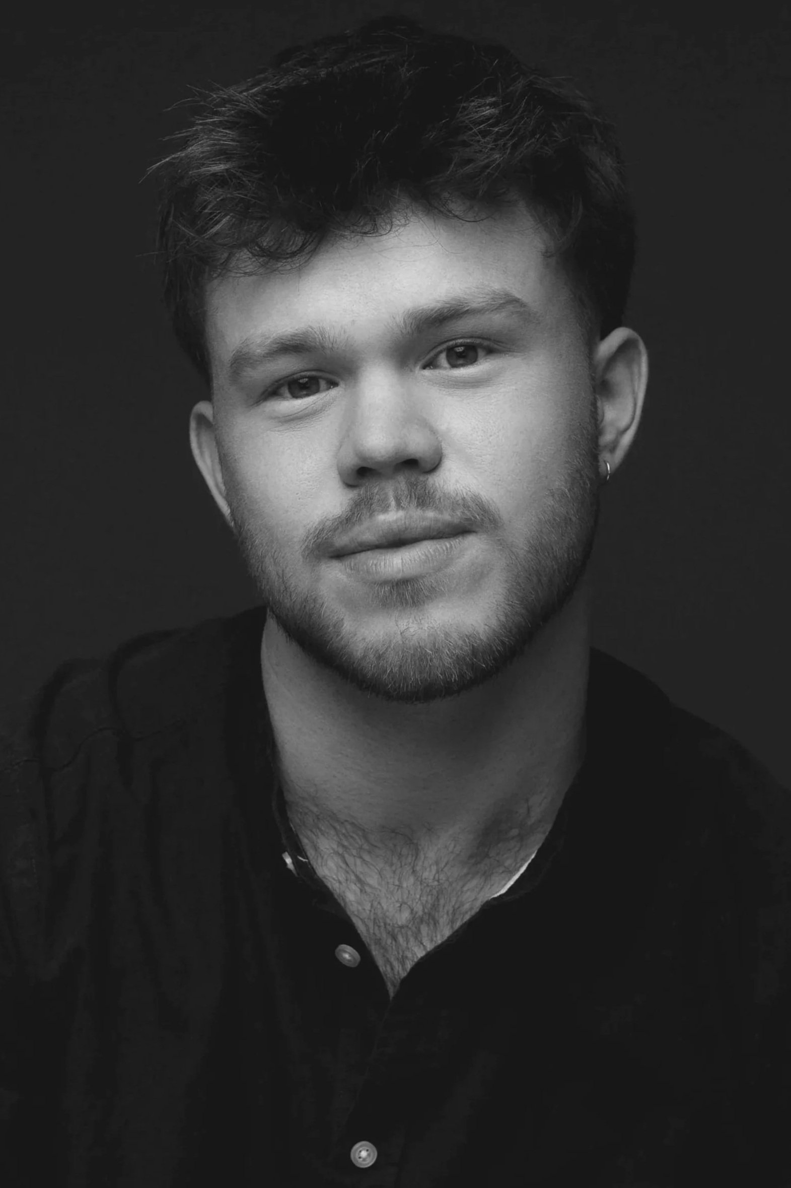 Black and white portrait of a young man with short, wavy hair, light beard, and wearing a dark shirt, against a plain dark background.