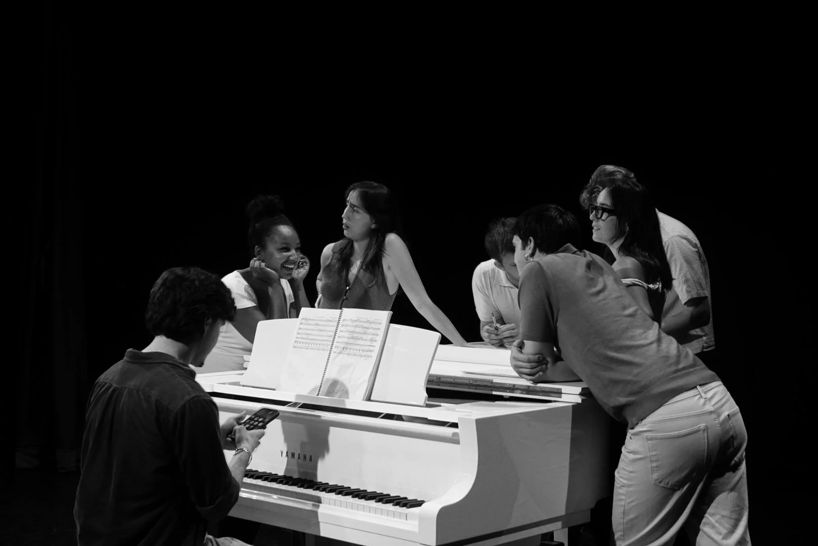Group of people gathered around a white Yamaha piano, some looking at sheet music, others engaged in conversation, in a dark room.