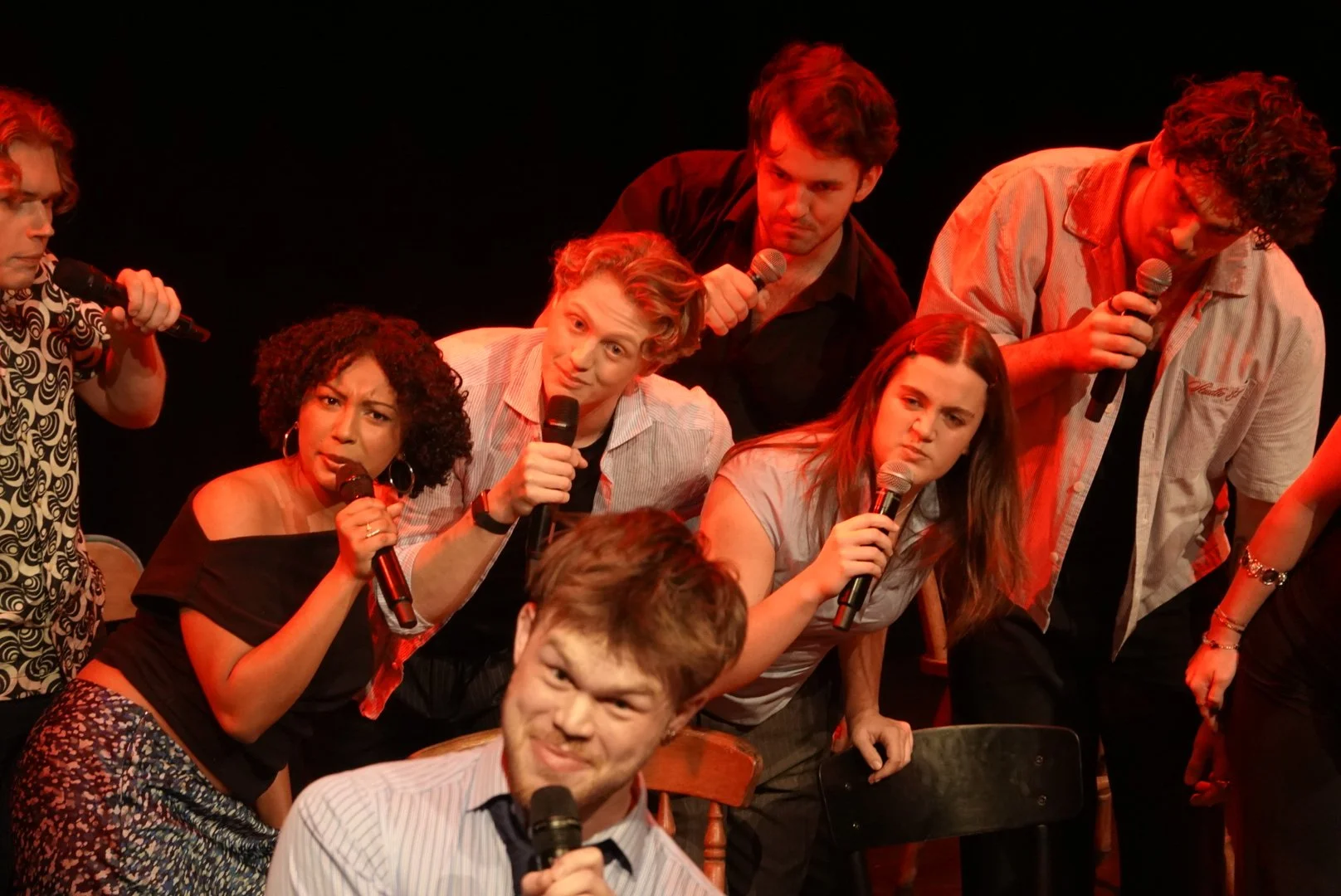 Group of people on stage holding microphones, some looking serious and some bewildered, with a dark background.